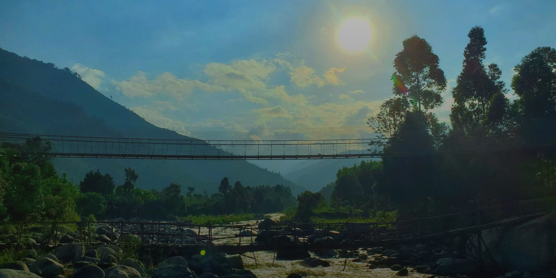 A scenic landscape featuring a mountain, trees, a river with rocks, and a suspension bridge under a bright sun with some clouds.