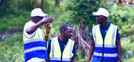 Three young men wearing high-visibility vests and white caps engaging in a discussion outdoors surrounded by greenery.
