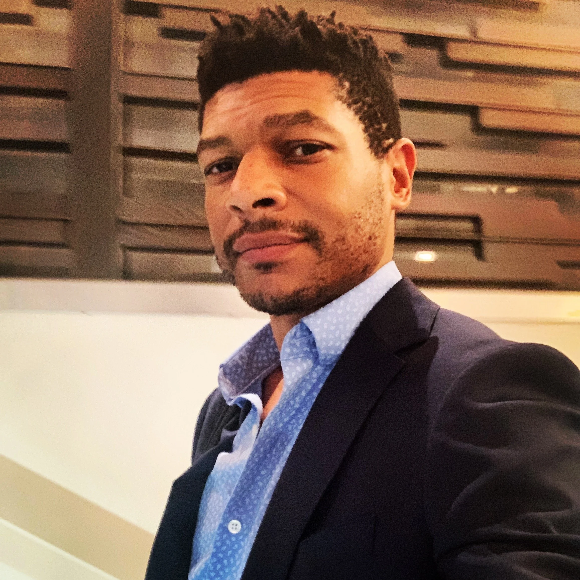 A young man with short curly hair, a mustache, and facial hair wearing a dark blazer and a light blue dress shirt with a subtle pattern, standing indoors in front of a wall with horizontal wooden slats.  This is CJ Harvey, the company principal