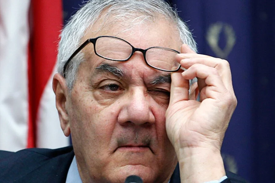 An older man with gray hair, holding glasses, winking and looking serious in front of an American flag.