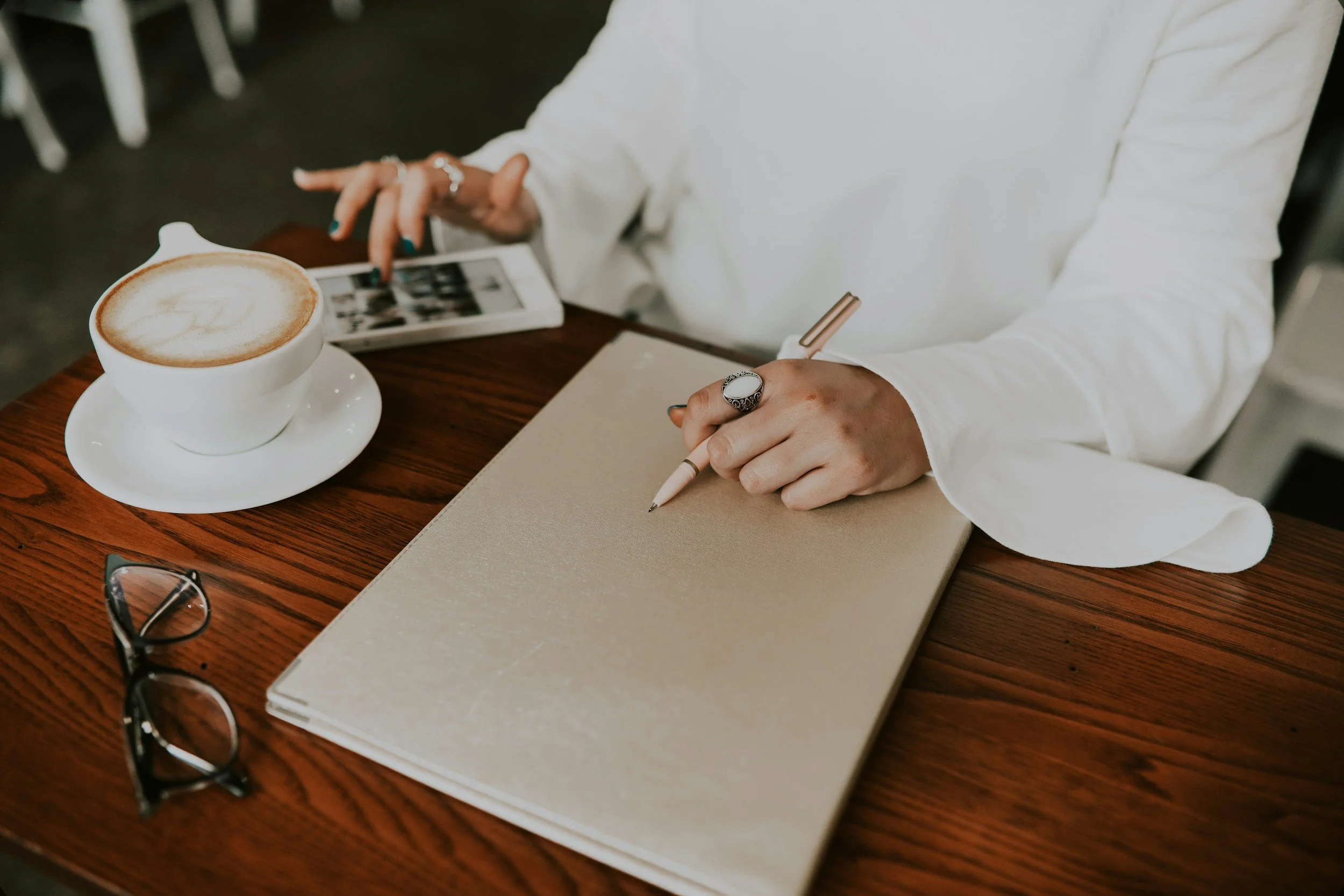 Person writing in a notebook at a wooden table with a cup of coffee, a smartphone, glasses, and rings.
