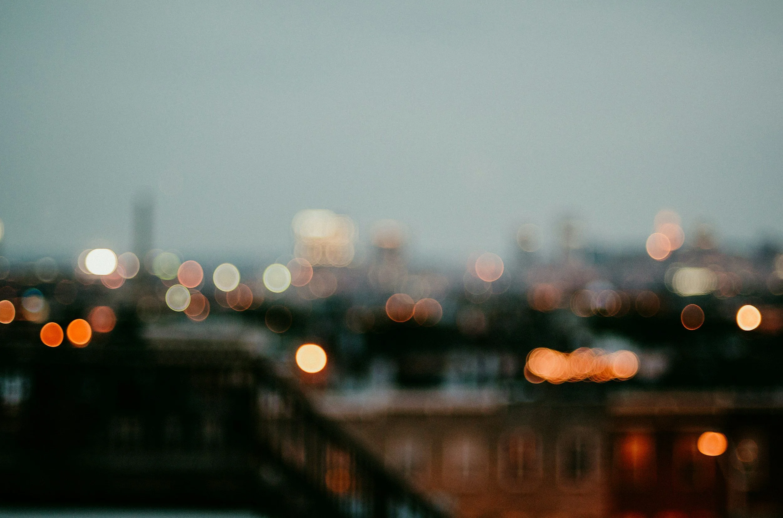 Blurred cityscape at dusk with out-of-focus multicolored lights and a dark silhouette of rooftops.