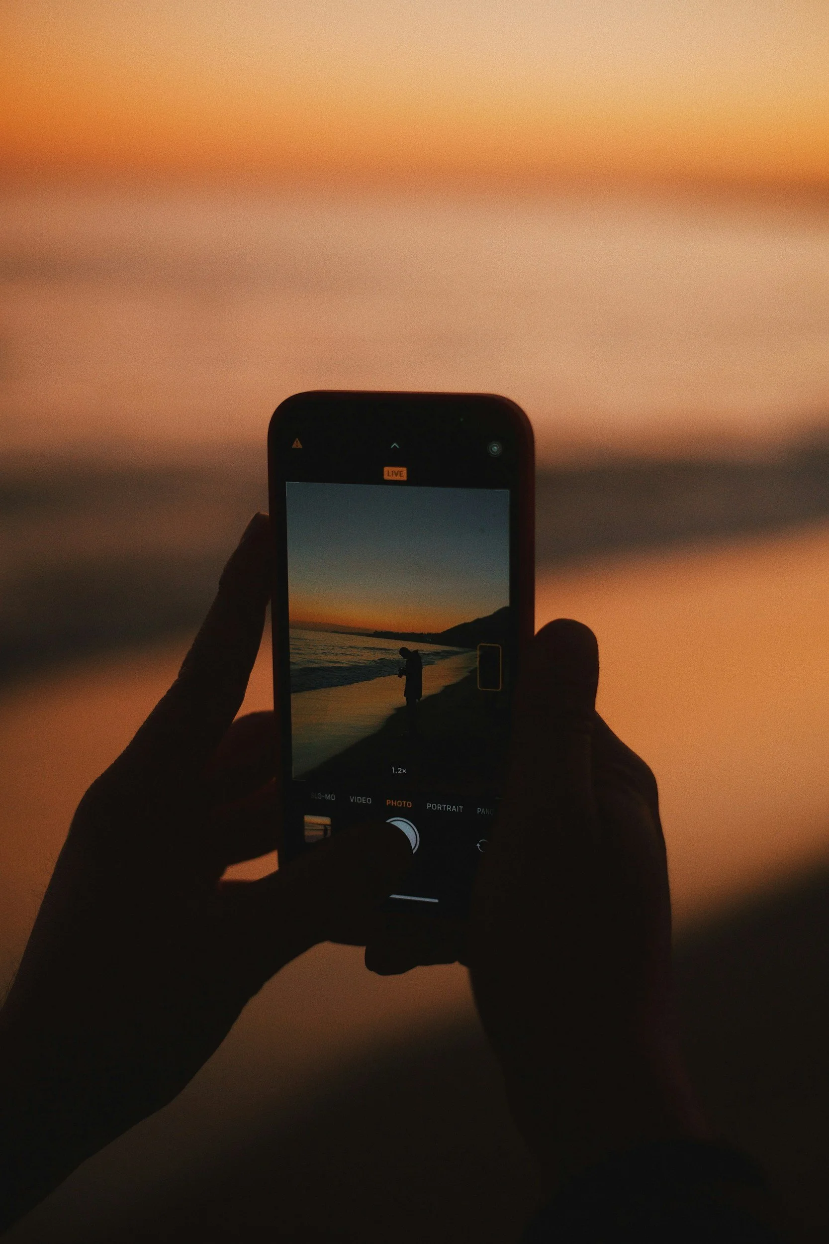 A person taking a photo of a sunset at the beach with their smartphone, showing the silhouette of another person near the shoreline.