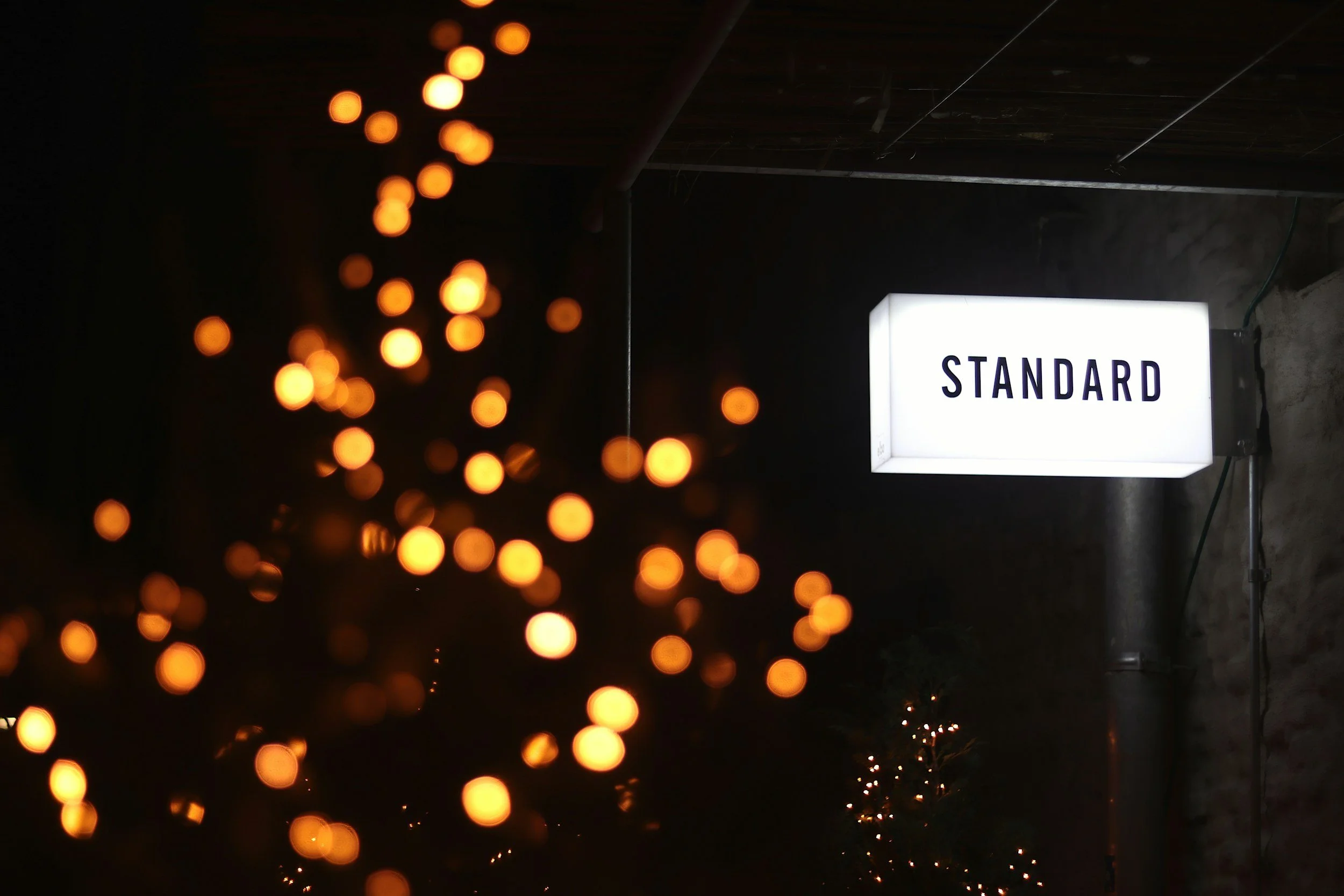 A white illuminated sign with the word 'STANDARD' hangs on a dark wall, with blurred orange lights in the foreground.