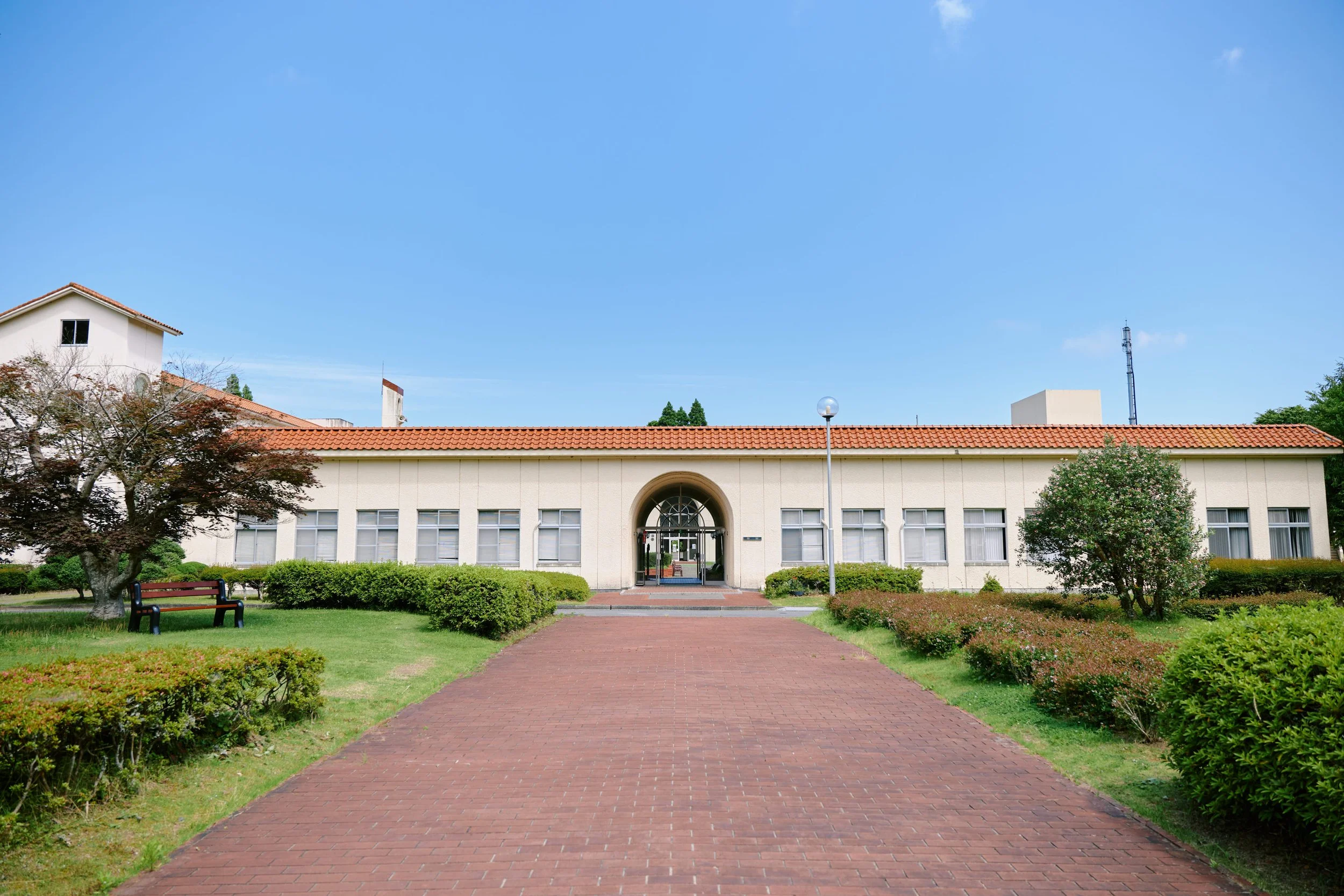 青空の下に建つ赤い屋根の白い建物。前には赤レンガの道路と緑の芝生と木。