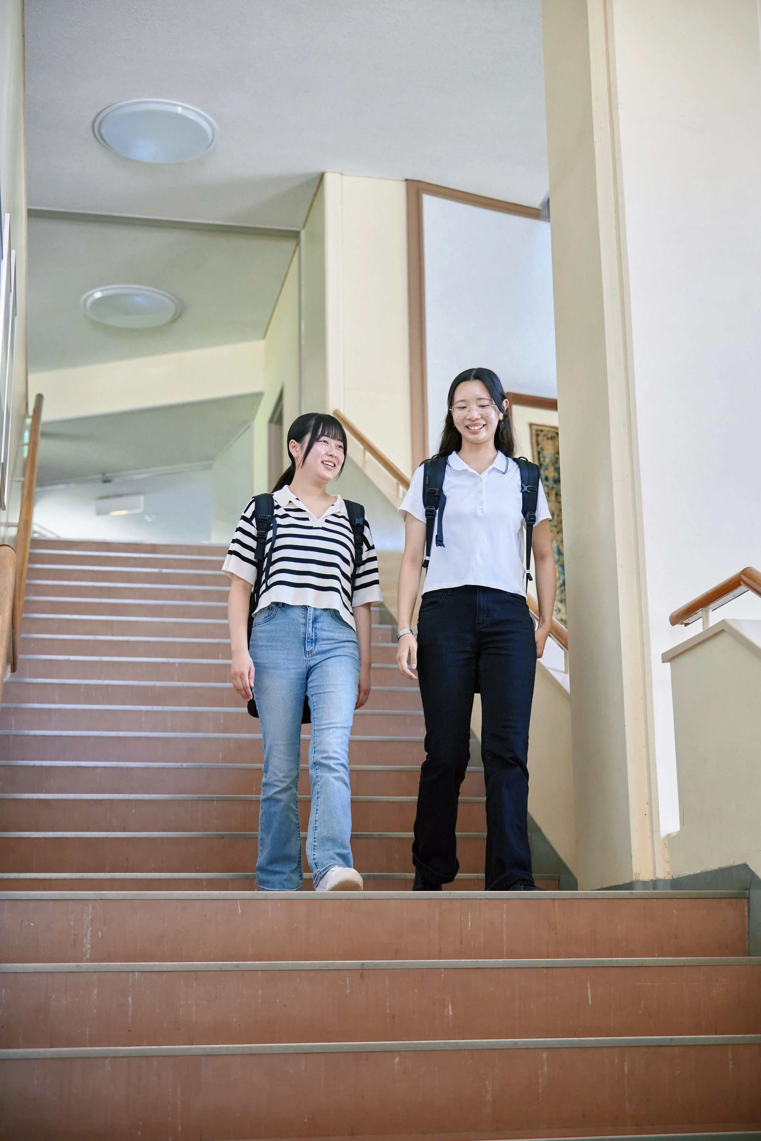 階段を下りる二人の若い女性。両者ともバックパックを背負い、笑顔で会話している。