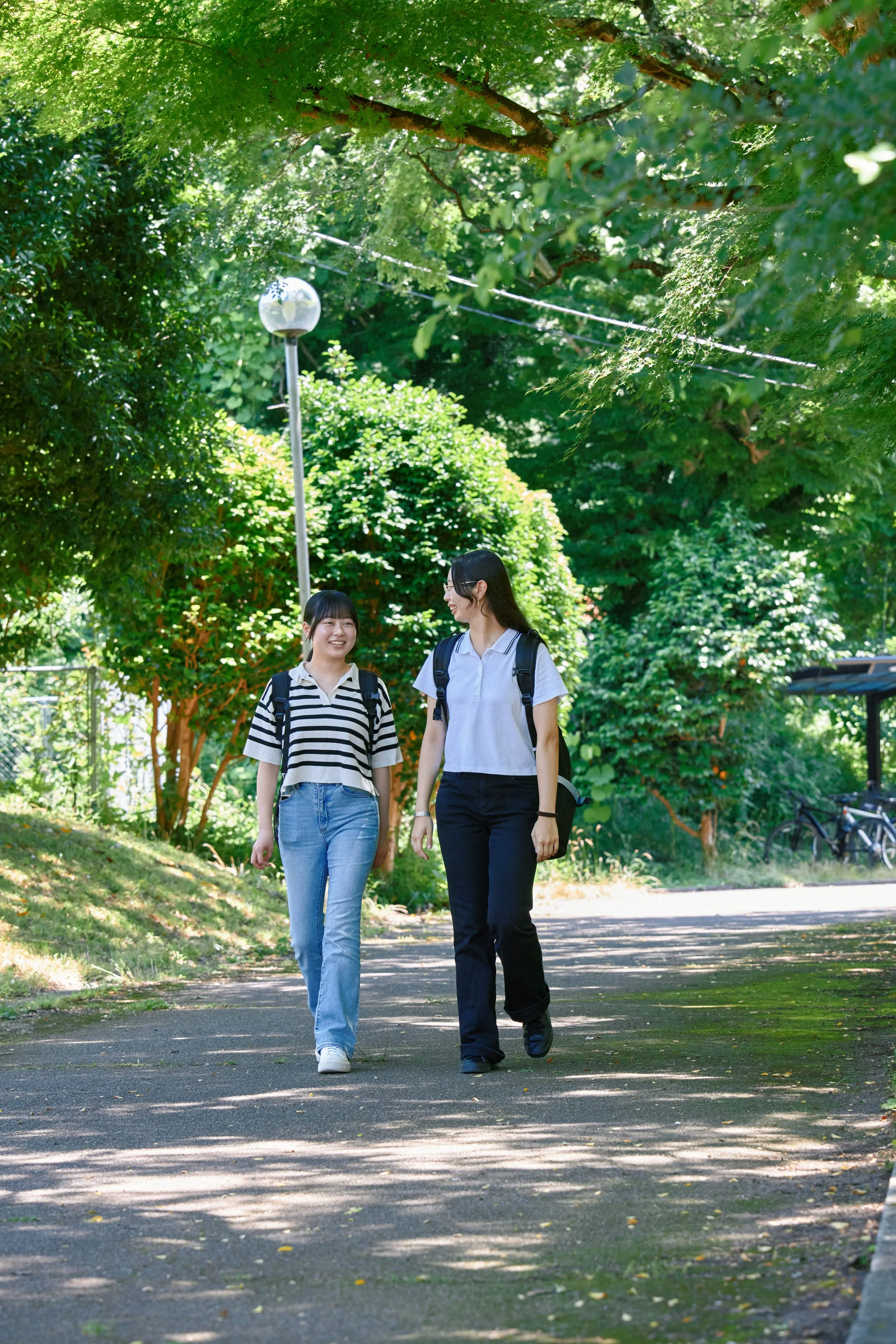 緑豊かな公園の歩道を歩く二人の若い女性。笑顔で話しながら、カジュアルな服とリュックサックを背負っている。日光が木の葉の間から差し込んでいる。