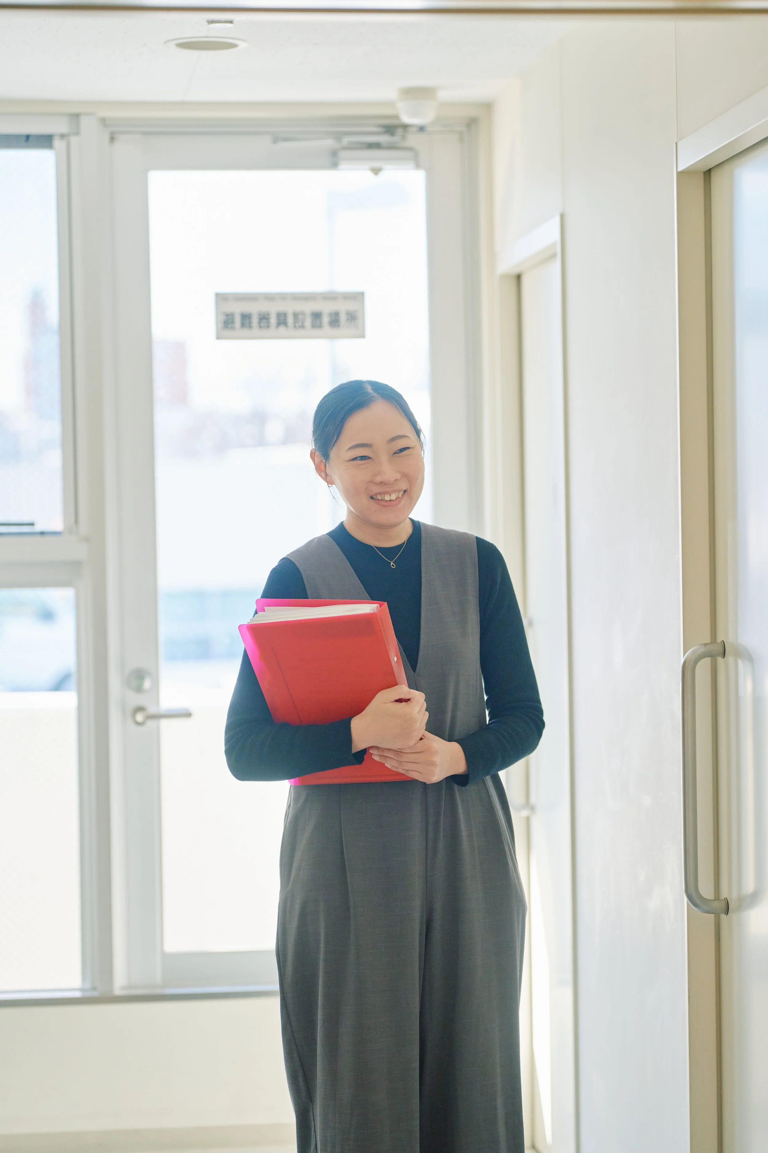 教室の中にいる笑顔の女性教師、手に赤いファイルを持っている