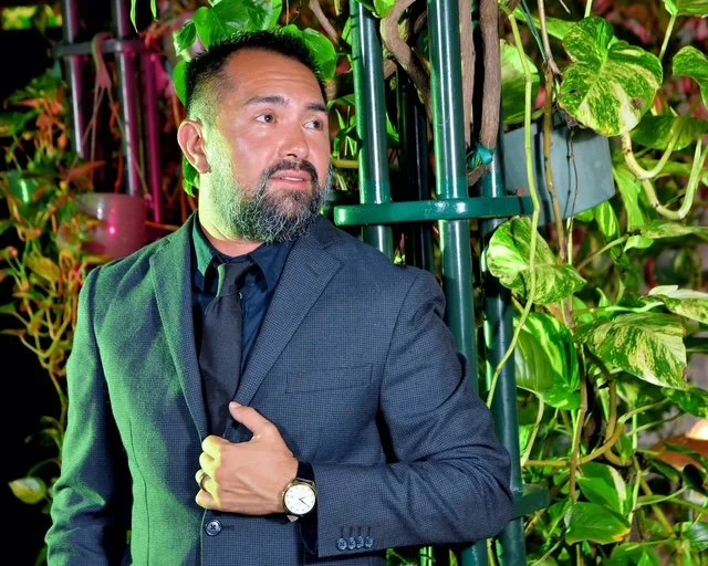A man in a dark suit and tie standing among green leafy plants with a metal structure in the background, outdoors at night.