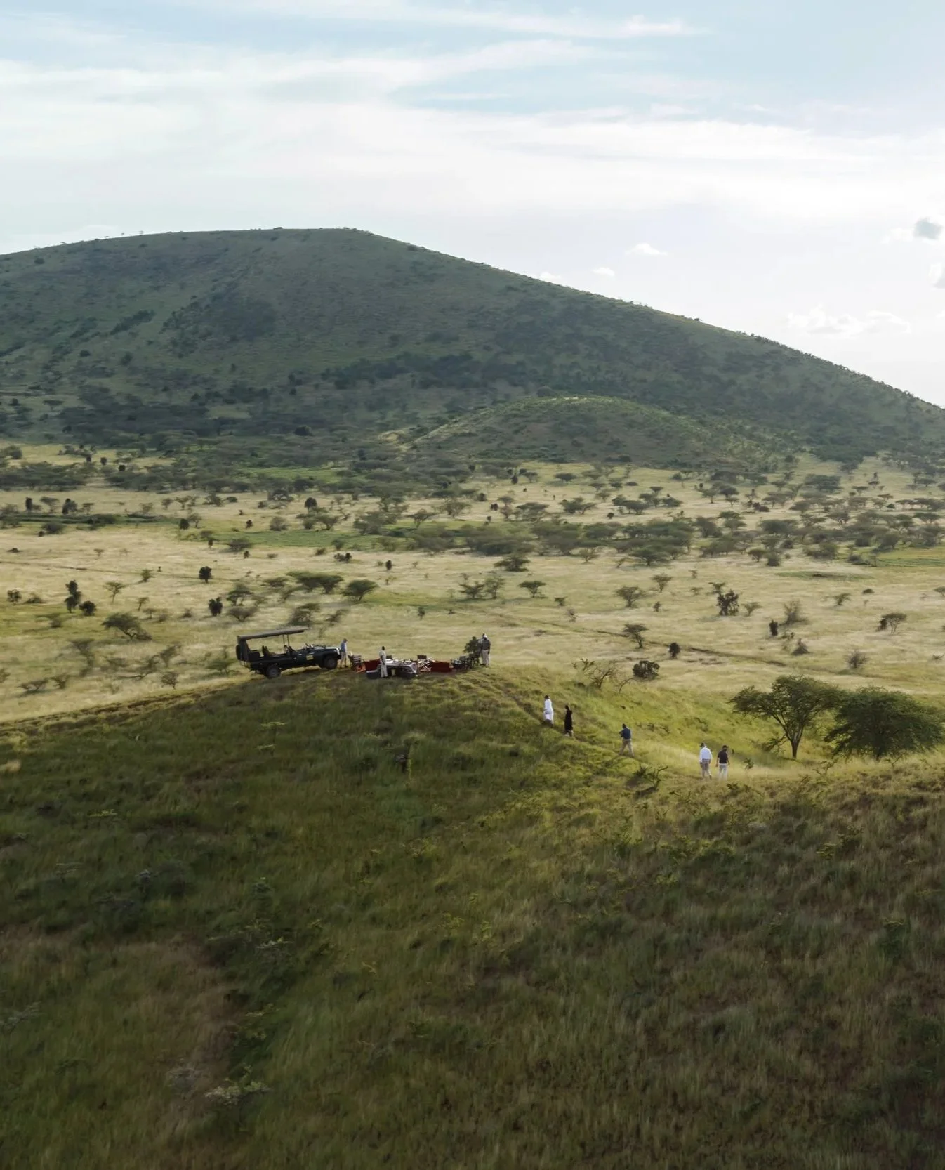 A group of people walking through a grassy landscape with small trees, a vehicle parked on a small hill, and a mountain in the background under a partly cloudy sky.