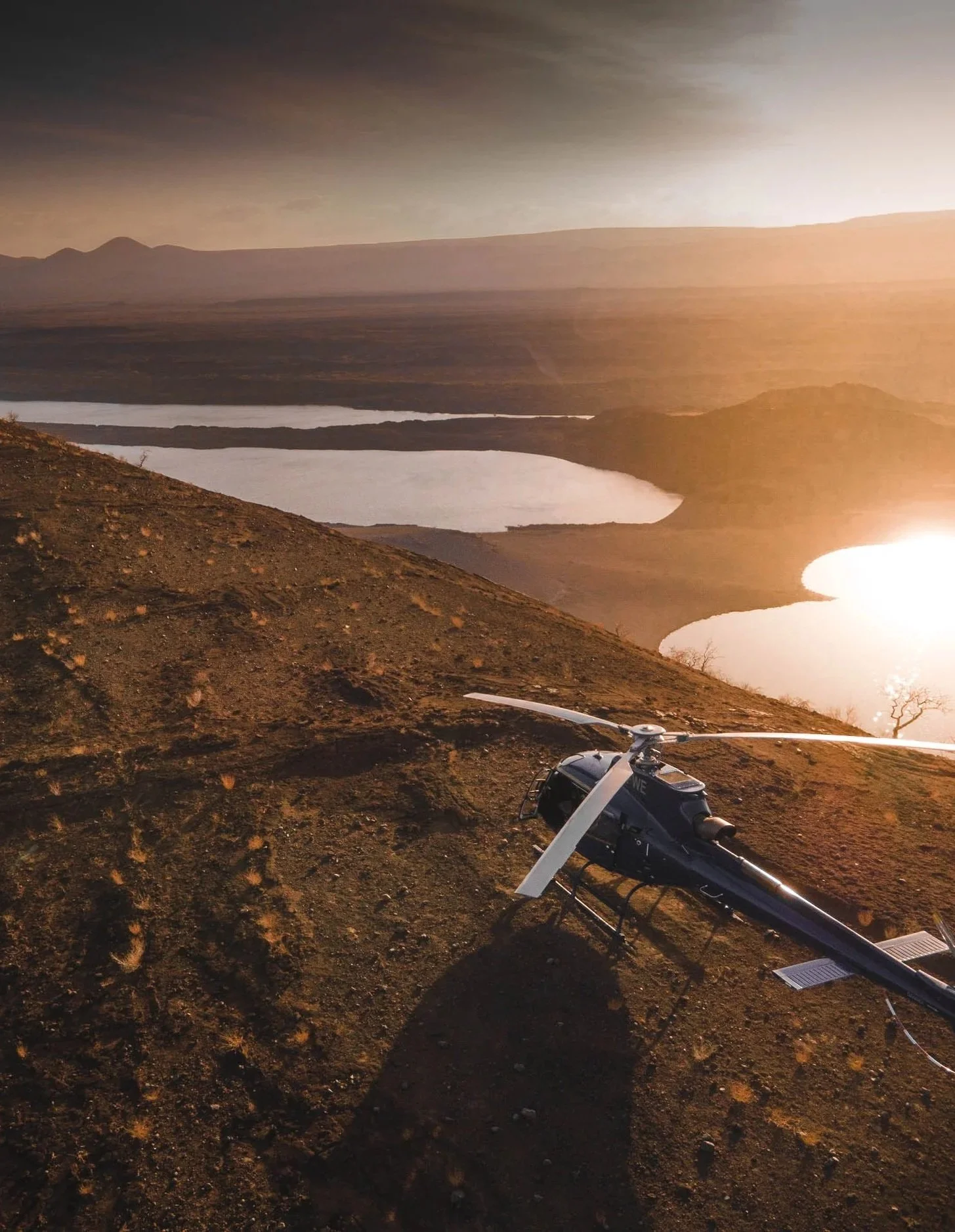 A black helicopter resting on a dirt hillside during sunset with lakes and mountains in the background.
