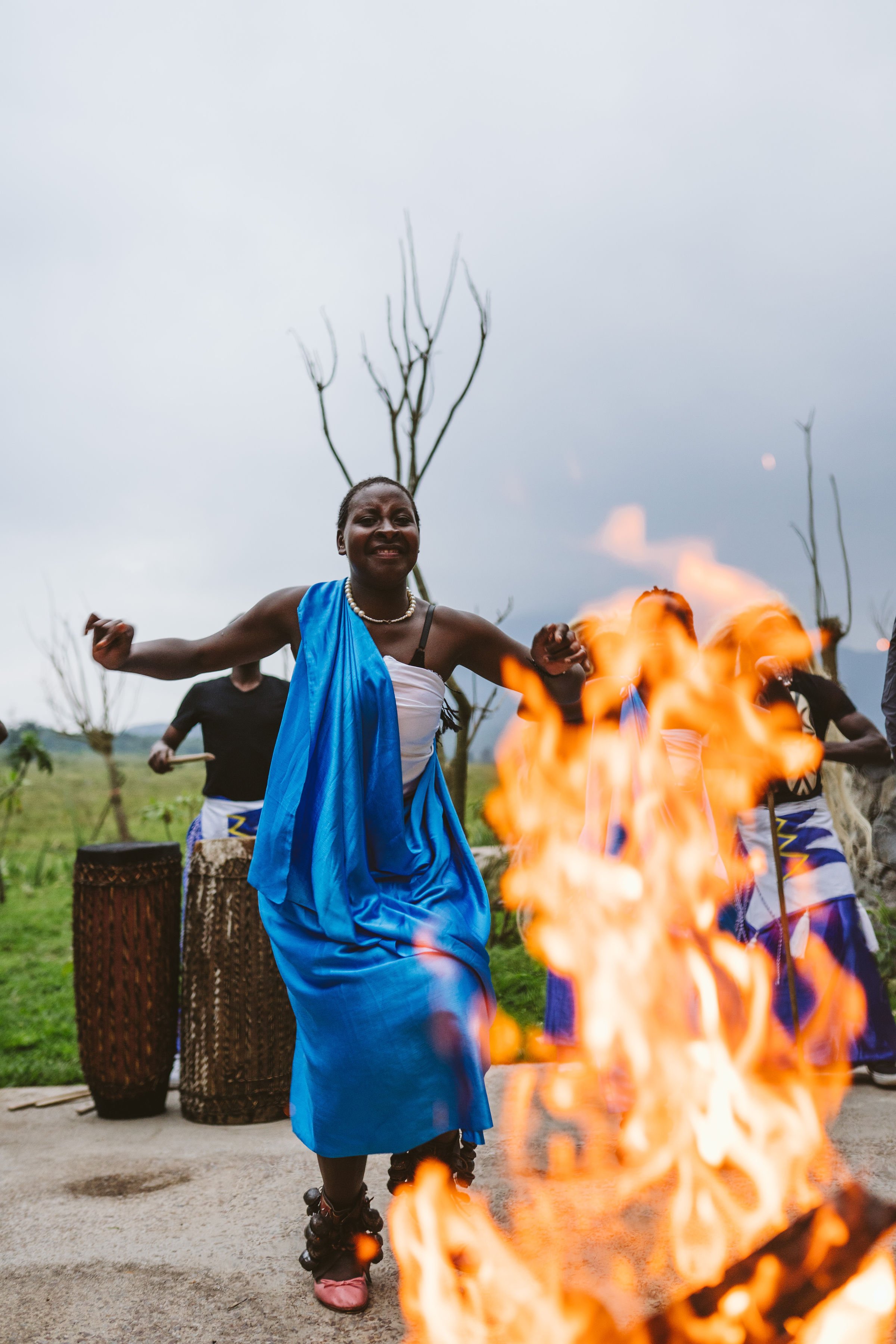 A woman in traditional attire dances around a fire as part of a cultural celebration, with others playing drums in the background outdoors.