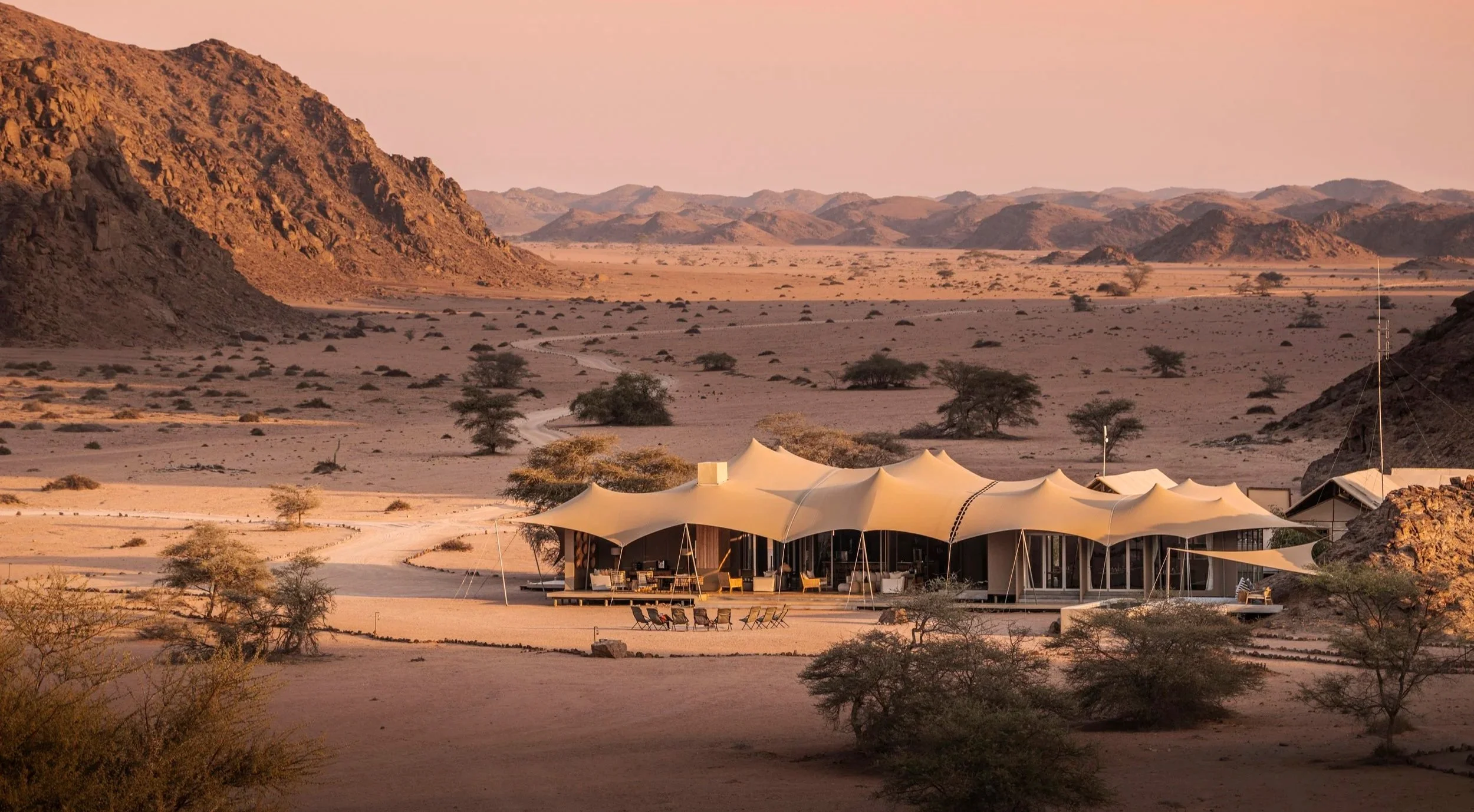 A luxury desert resort with a cream-colored tensile tent-shelter structure, surrounded by sparse desert shrubs and mountains in the background during sunset or sunrise.