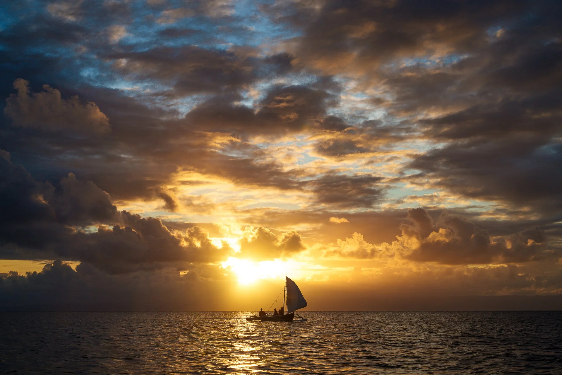 A sailboat with two people aboard on calm ocean water during sunset, with a partly cloudy sky and golden sunlight.