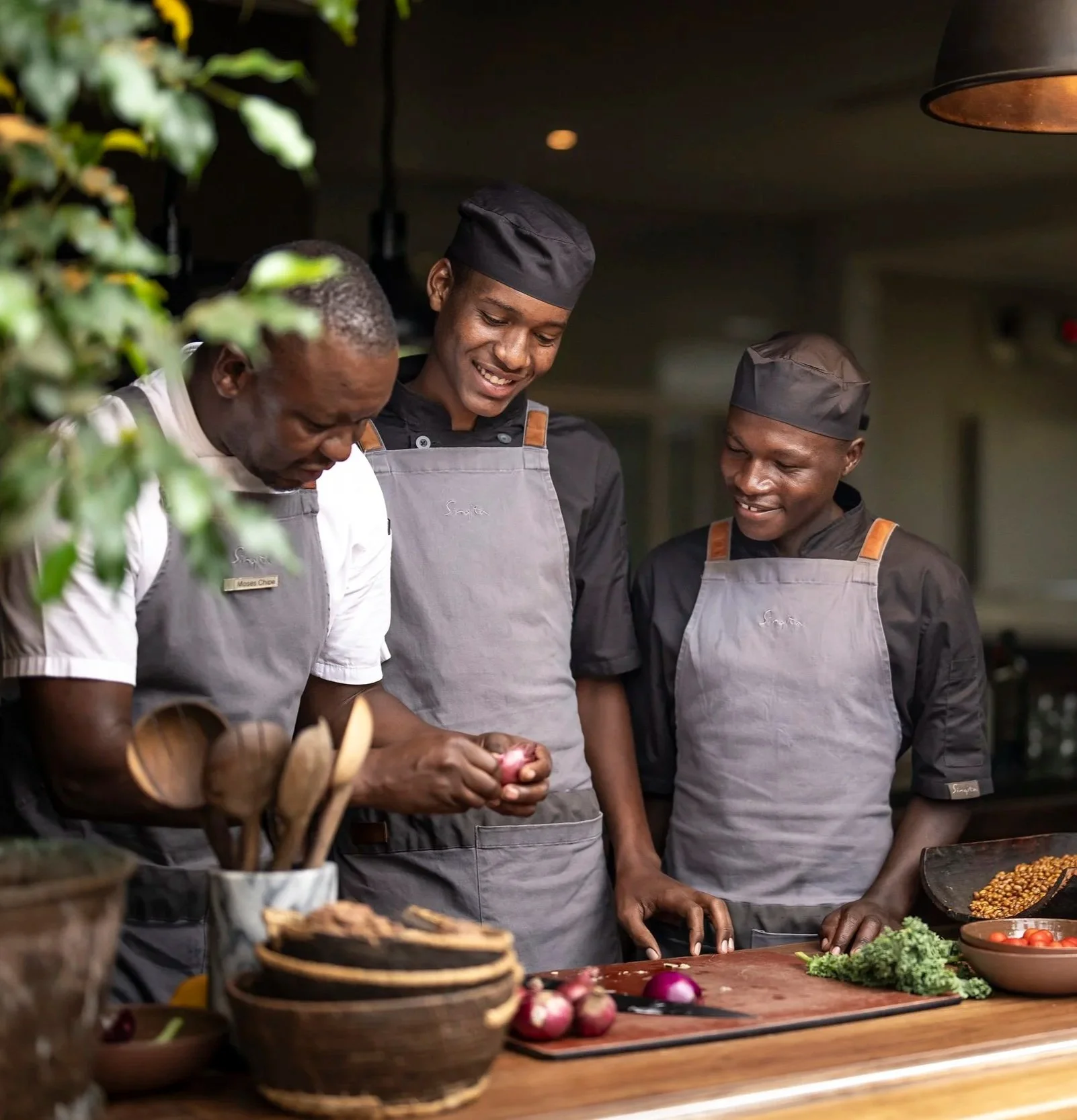 Three male chefs in black uniforms and gray aprons preparing vegetables and ingredients in a professional kitchen, smiling and working together.