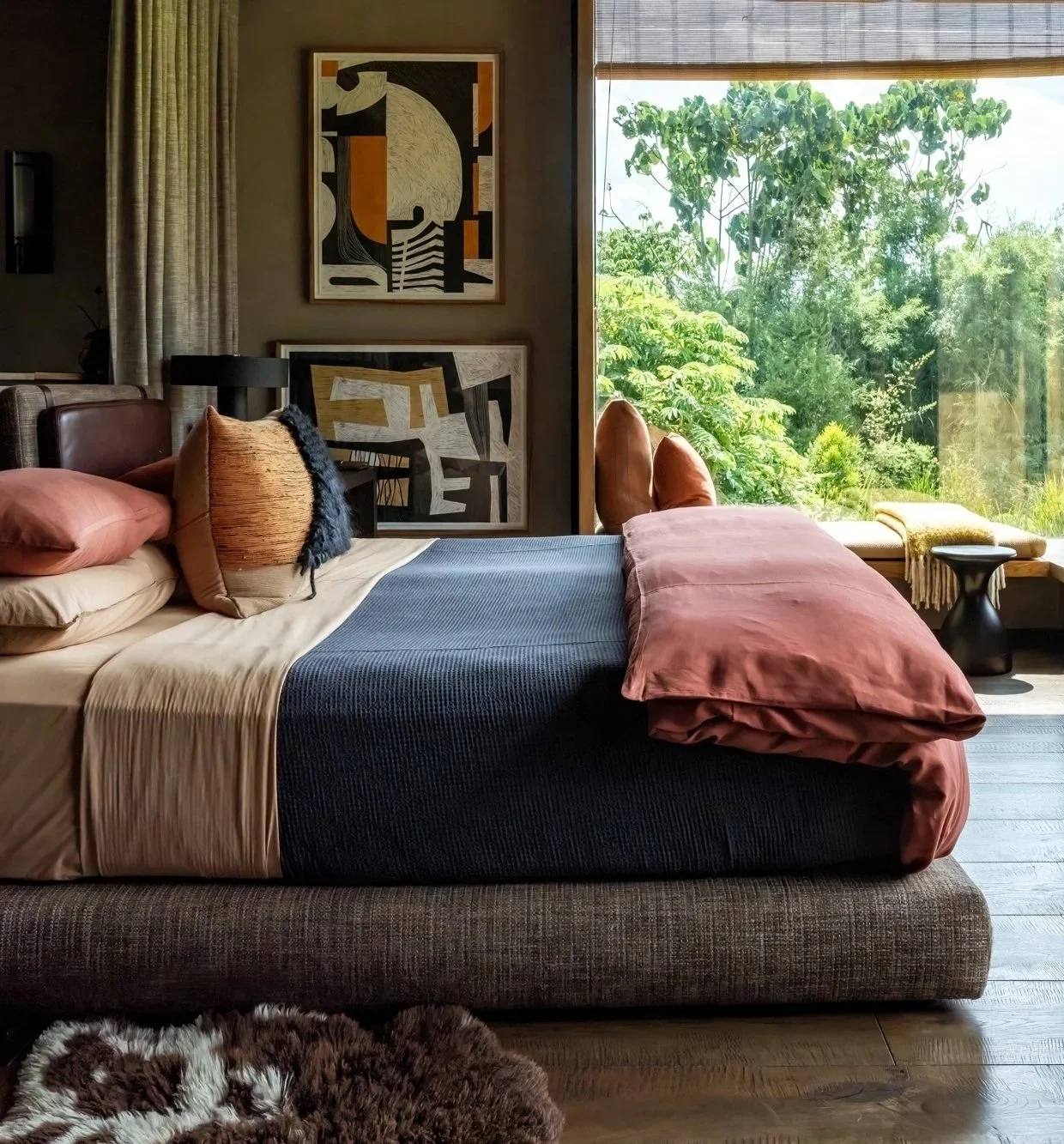 A cozy bedroom with a bed covered in pink and dark blue bedding, decorative pillows, and a large window showing green trees outside.