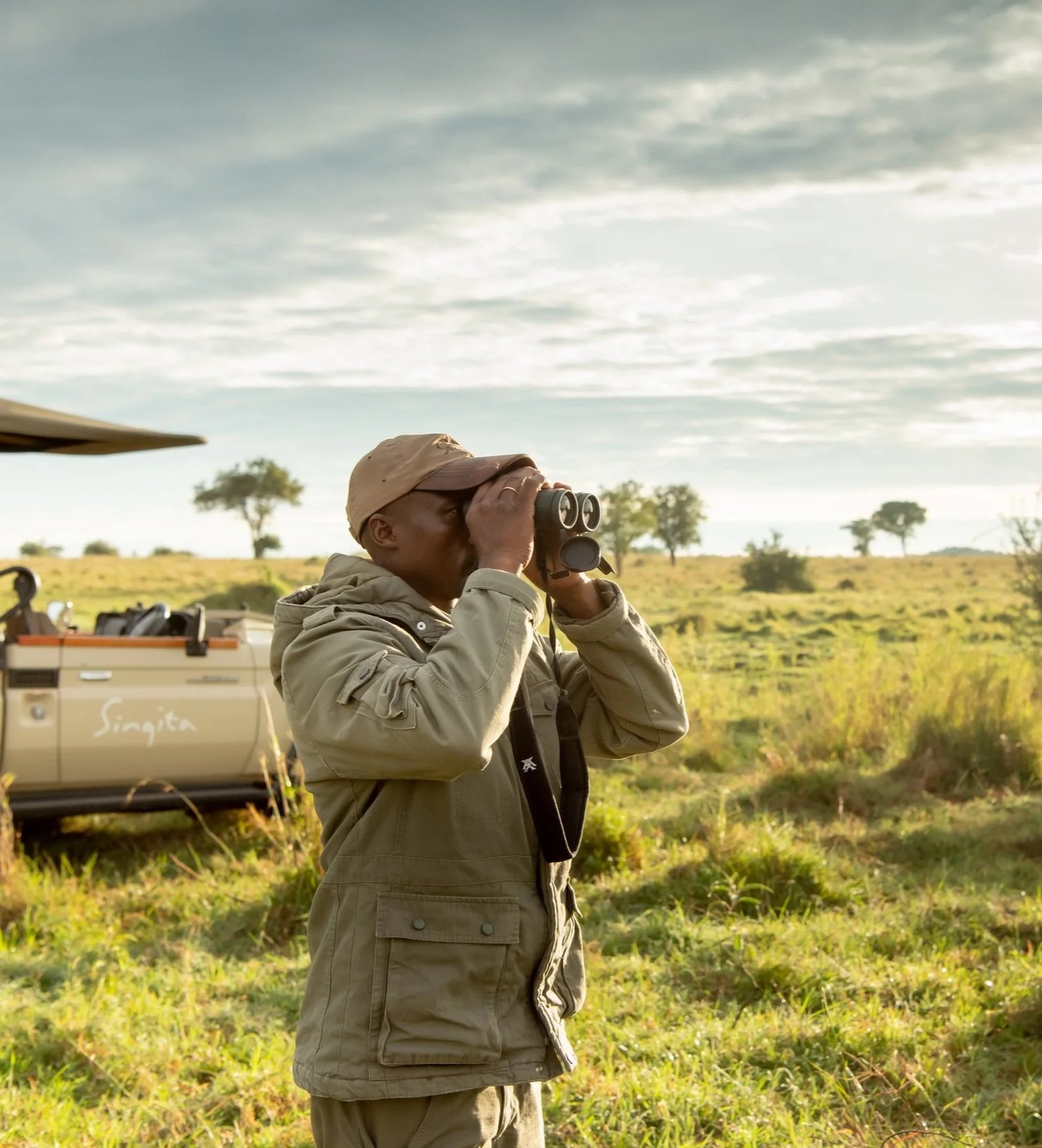 A man wearing outdoor clothing and a hat looking through binoculars in a grassy savanna landscape with trees, during daytime.