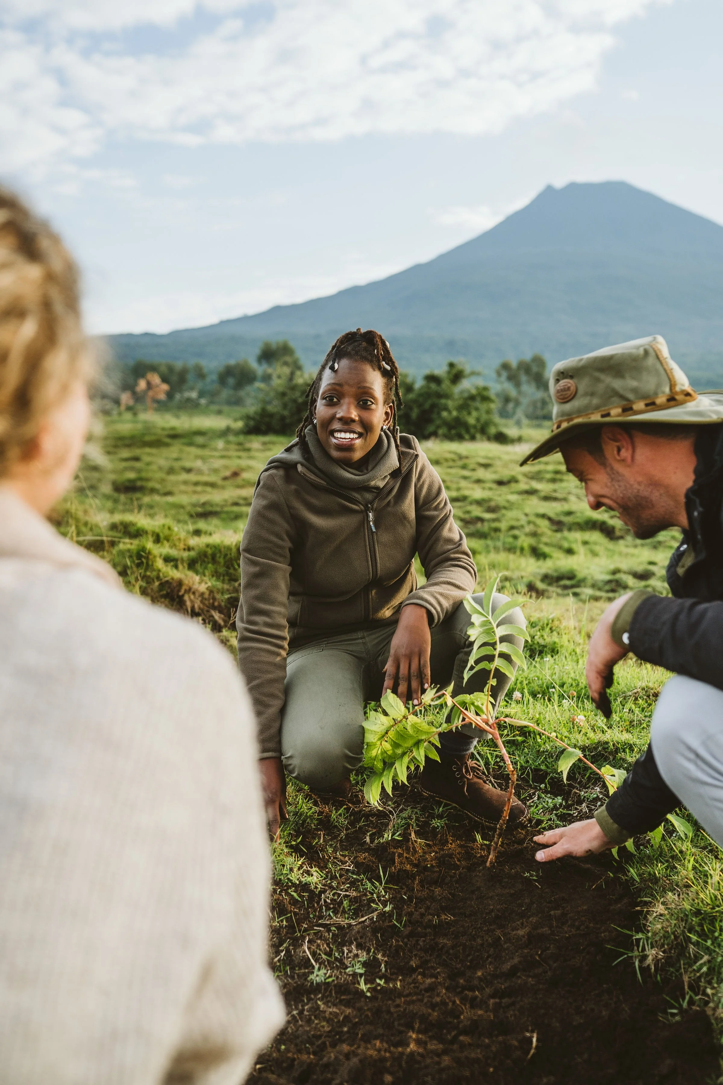 Three people planting a tree in a lush green field with mountains in the background. One woman is smiling, kneeling, and holding the young tree, while a man and another person are engaged in planting it.
