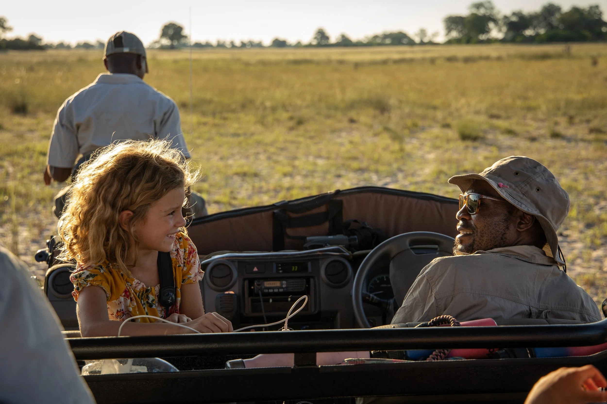 A young girl and an older man are seated inside an open vehicle, smiling and talking, with a person walking away in the background across a field at sunset.