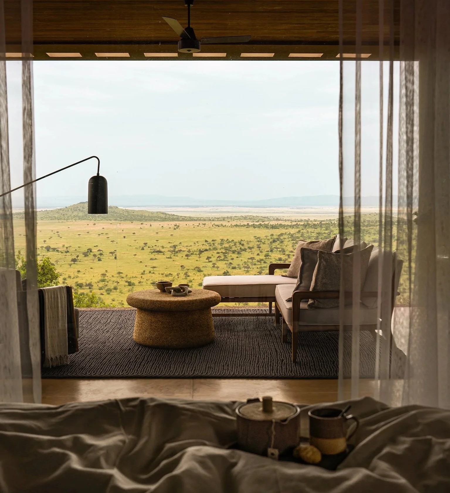 View of a savannah landscape from a room with a balcony, with outdoor furniture including a beige cushioned sofa and a round coffee table, and a ceiling fan overhead.