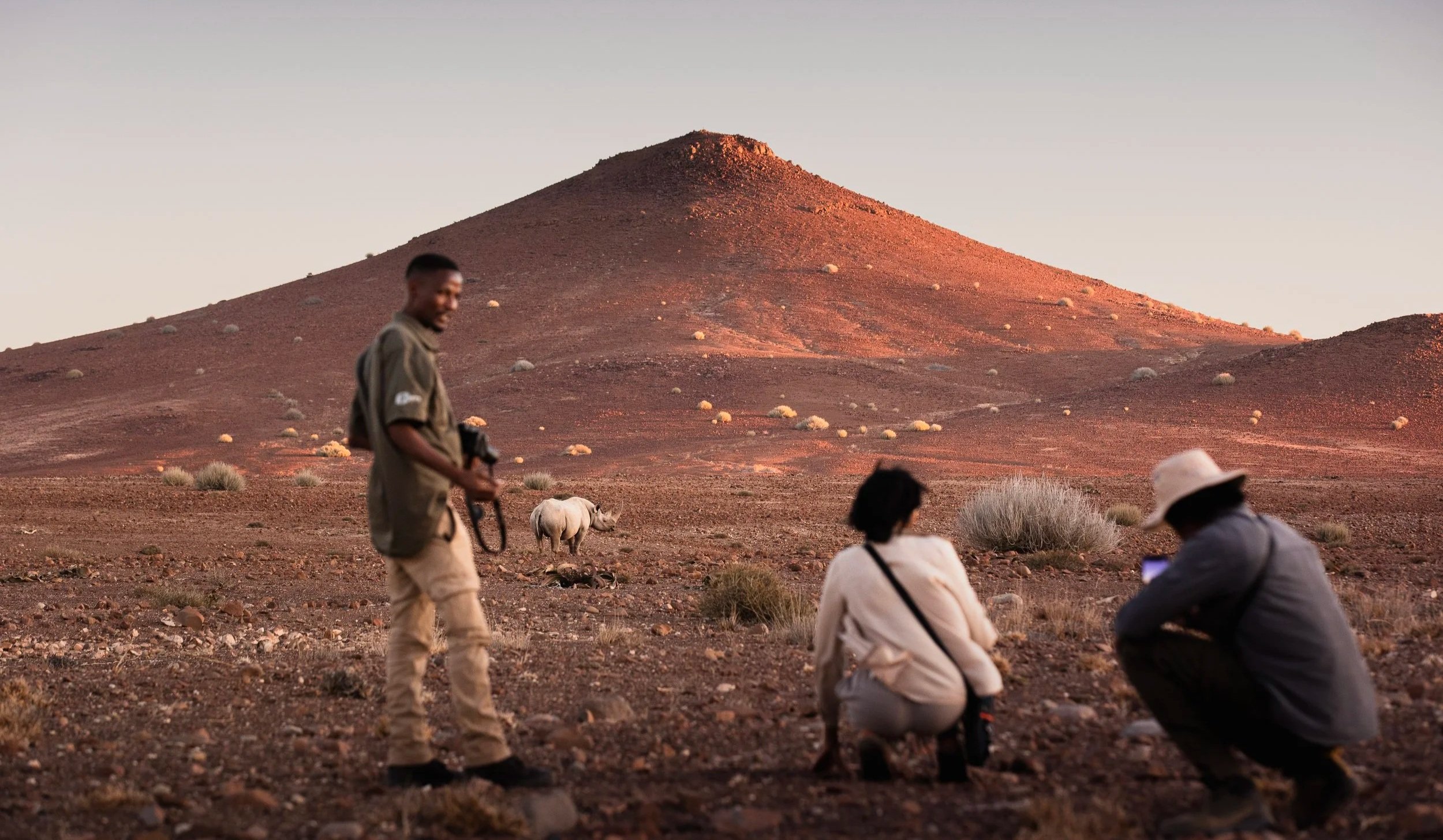 Three people in a desert landscape with a mountain in the background; one man is standing with a camera, and two women are crouching, one of whom is using a smartphone. A rhinoceros is seen in the distance near some shrubs.