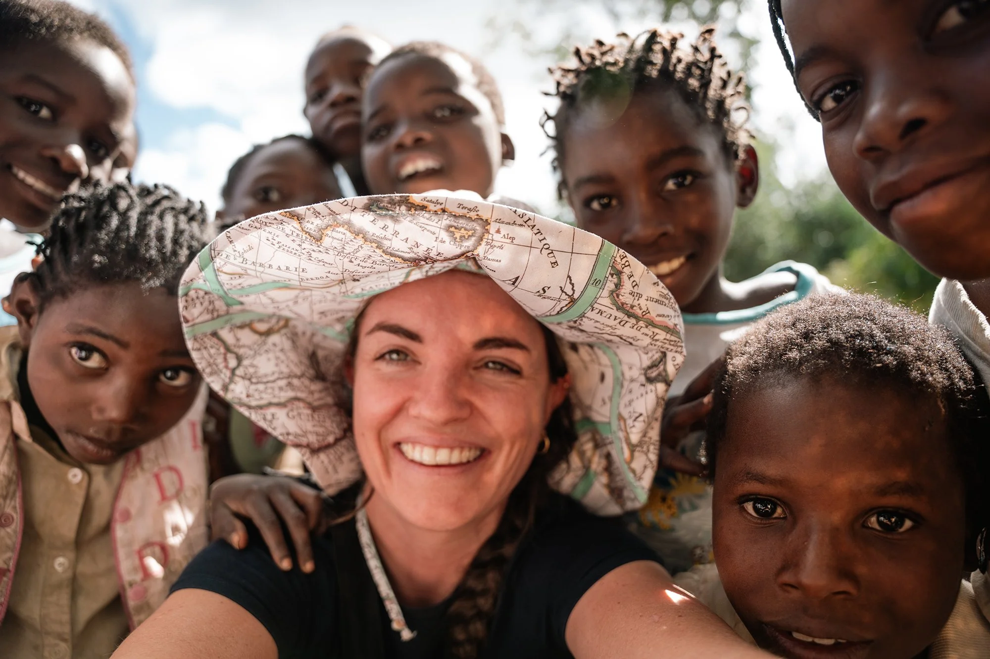 A woman wearing a wide-brimmed hat with a map pattern, smiling, taking a selfie with a group of children around her outdoors on a sunny day.