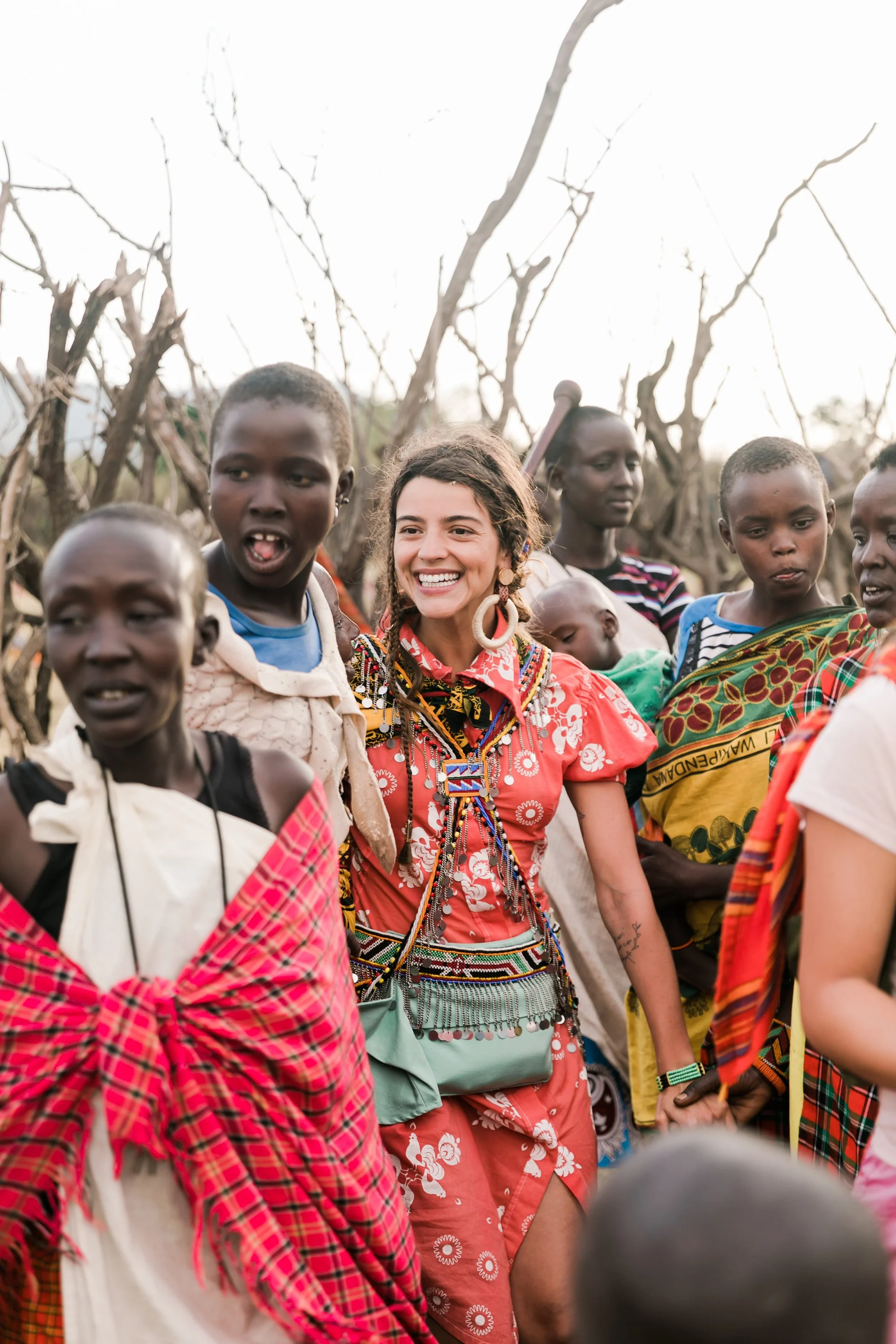A group of people, including a smiling woman wearing vibrant traditional clothing and jewelry, gathering outdoors among leafless trees, with some people looking in different directions.