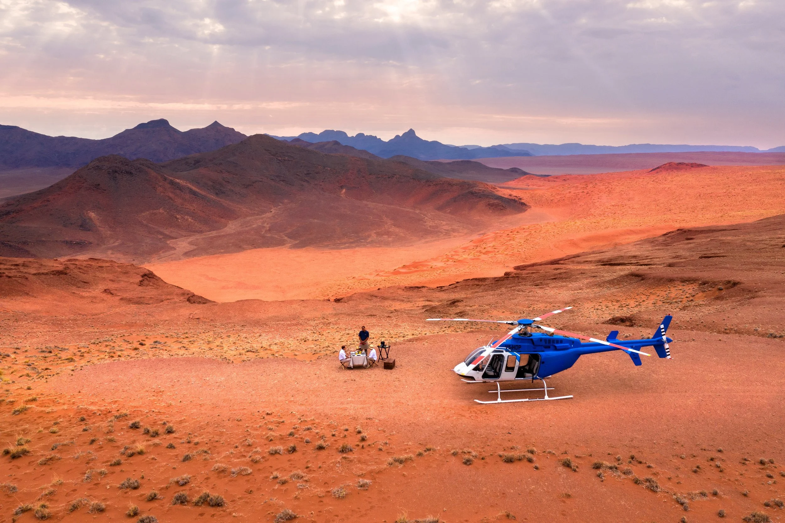 A helicopter in a desert landscape with mountains in the background, with three people sitting around a table and one person standing nearby.