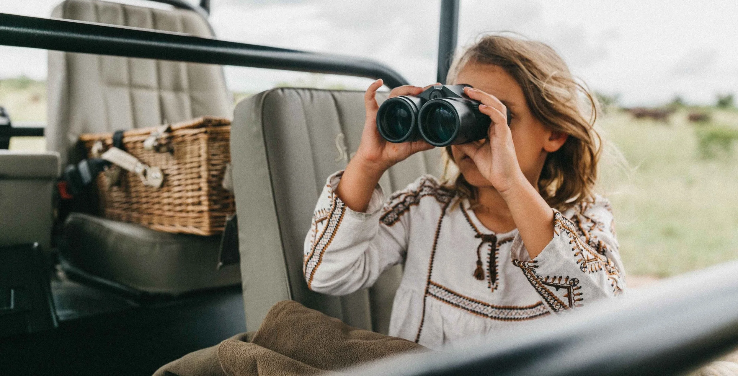 A young girl with curly hair wearing a white embroidered shirt looking through binoculars inside a vehicle with a grassy landscape outside.