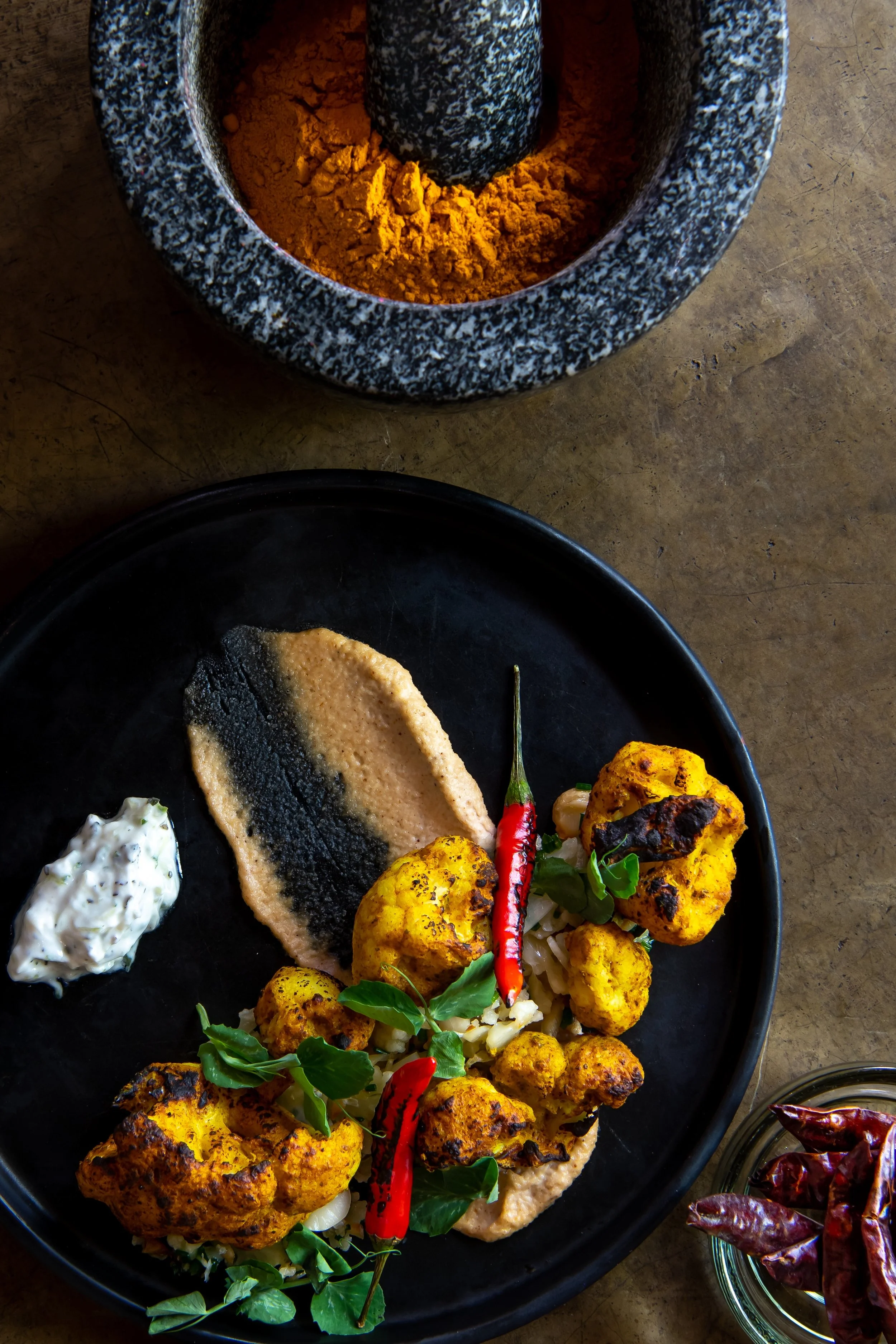 Plate of roasted cauliflower served with a chili pepper, a dollop of sauce, and a smear of hummus, with dried red chili peppers in a small bowl nearby, and a mortar with turmeric powder in the background.