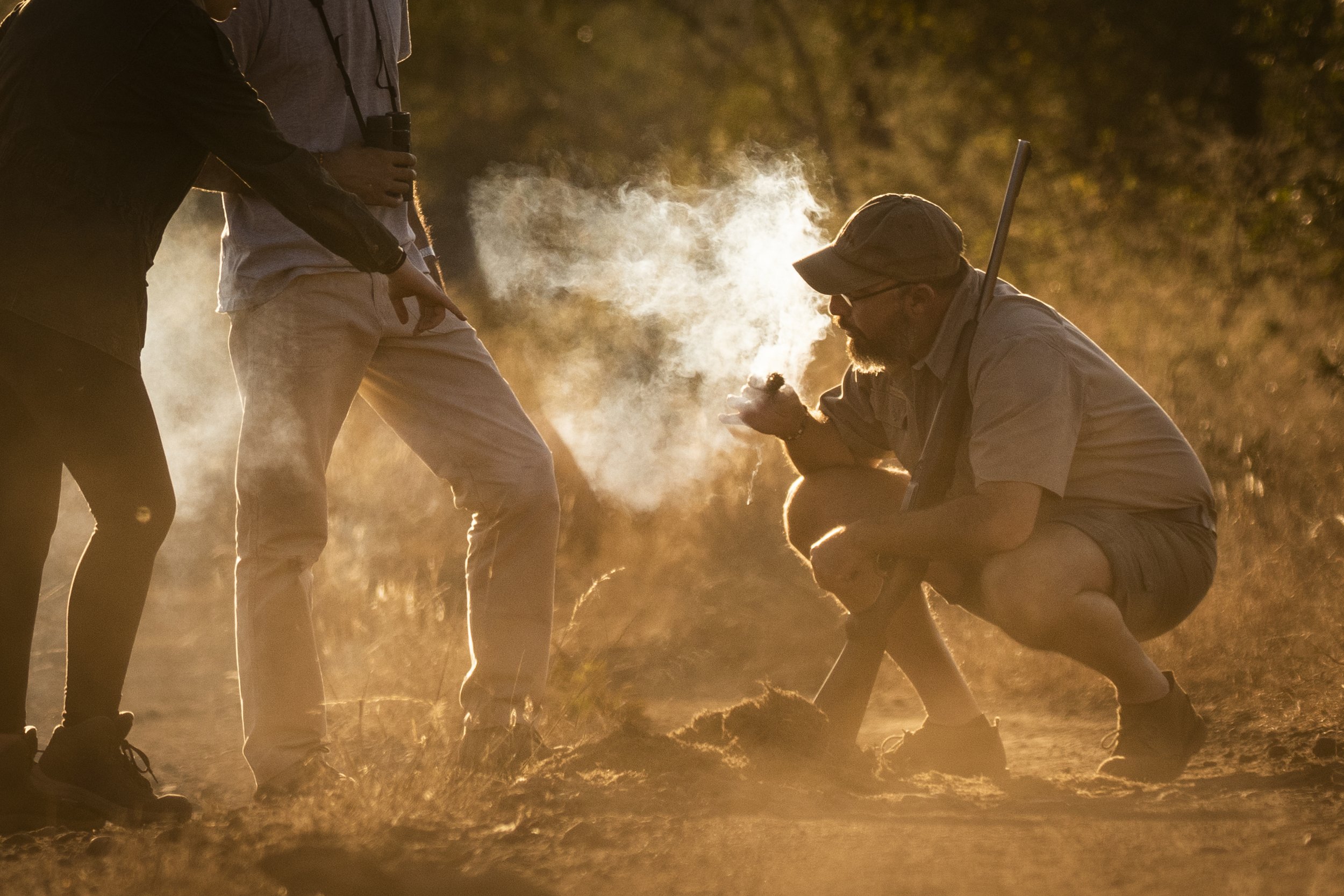 A man squatting and smoking while holding a stick, with two other people standing nearby, in an outdoor setting during sunset.