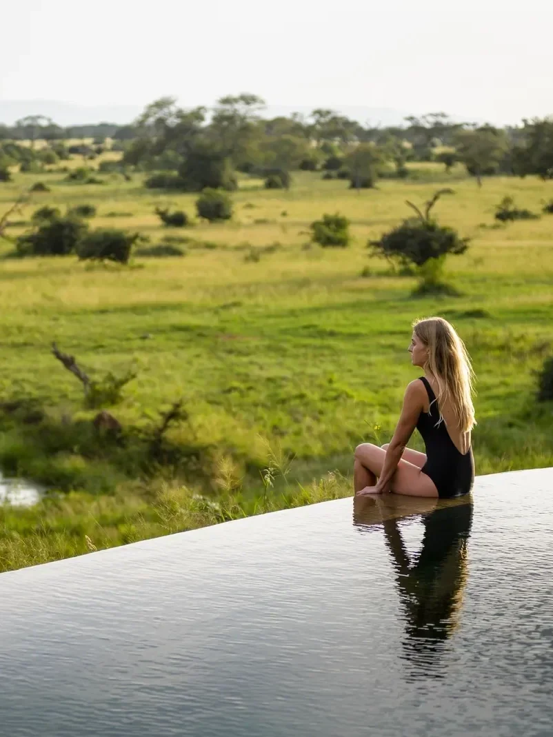 A woman in a black swimsuit sitting cross-legged on the edge of an infinity pool, overlooking a green grassy landscape with scattered trees.