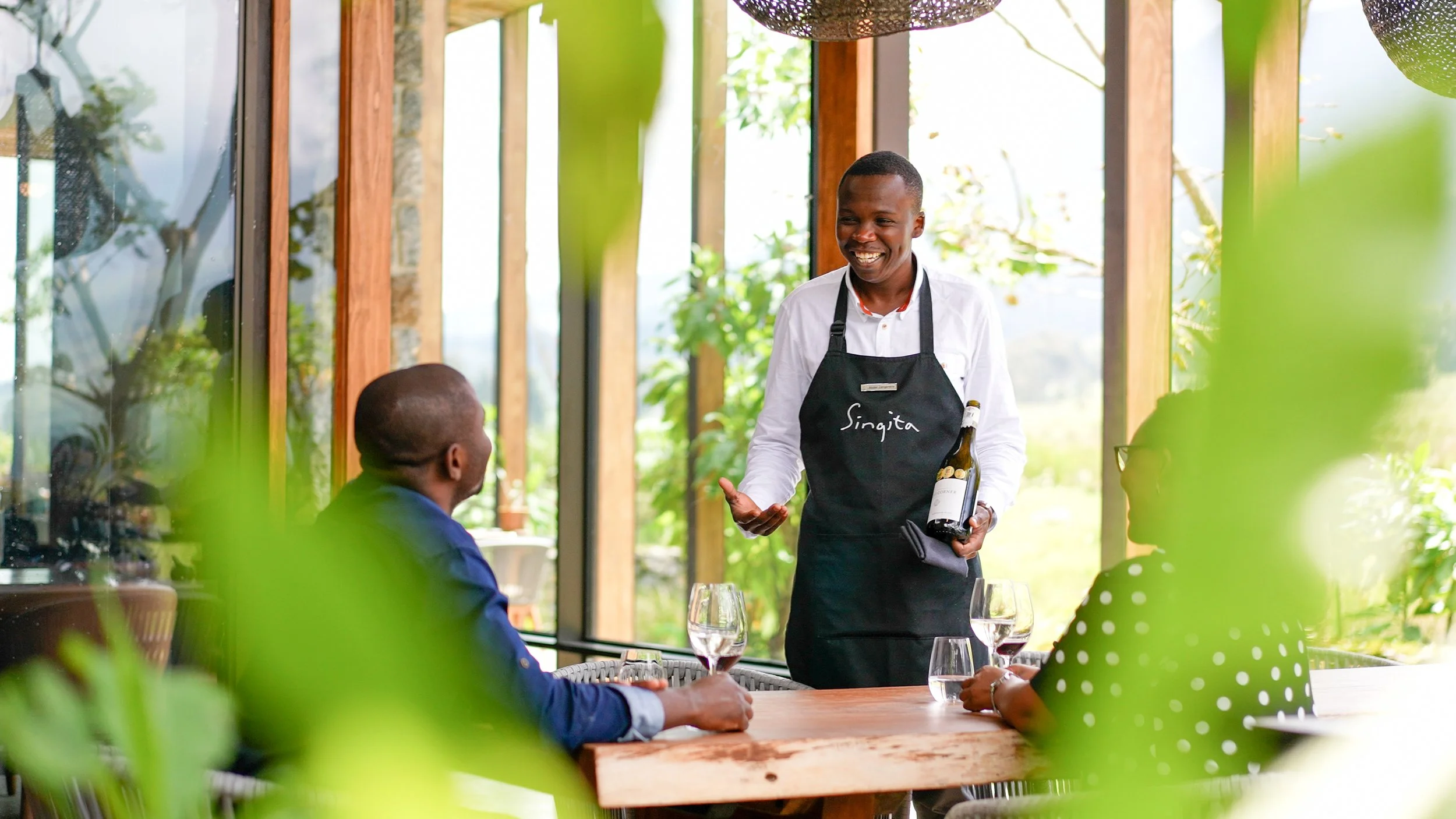 A smiling waiter holding a bottle of wine serving two seated customers at a table in a bright, lush restaurant with large windows and greenery outside.