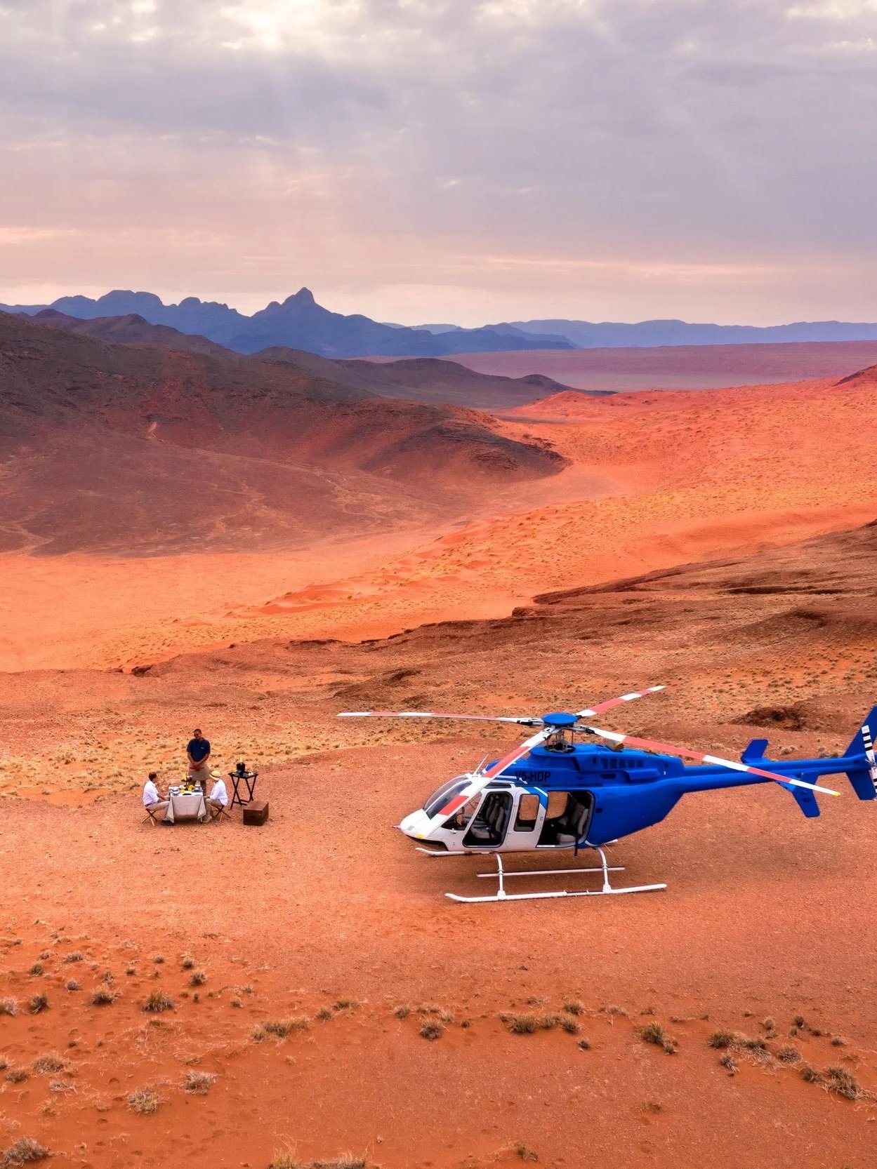 A blue helicopter parked on a desert plain with a table and chairs set up nearby, and two people sitting at the table. Mountainous landscape in the background during sunset or sunrise.