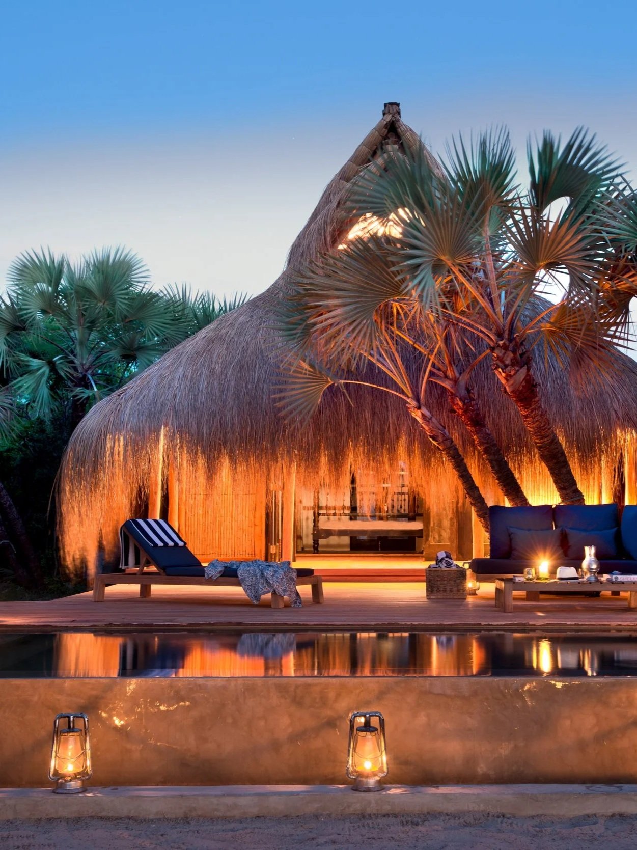 Tropical bungalow with thatched roof, surrounded by palm trees, lit by warm lights at dusk, with a pool and outdoor furniture.