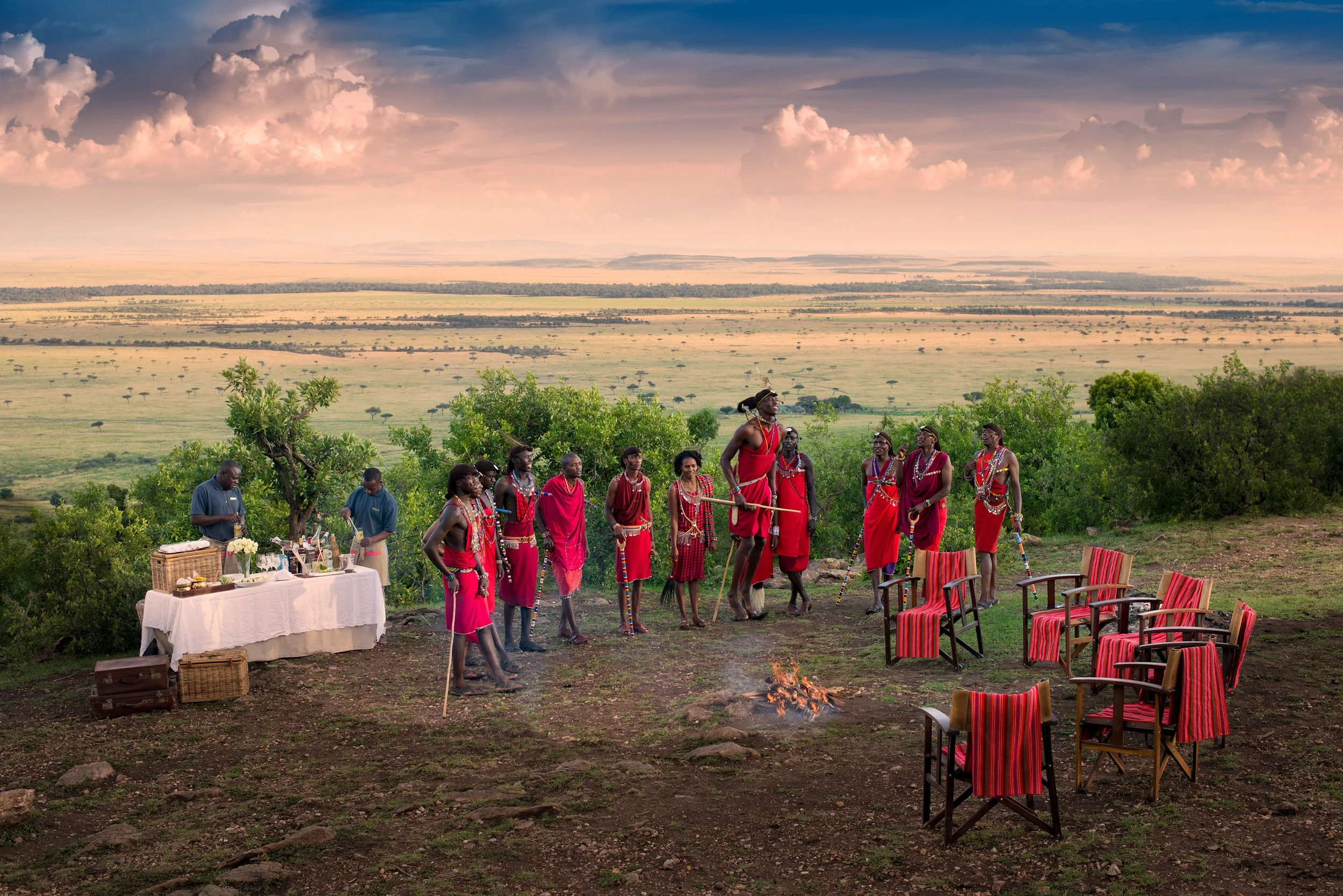 A traditional outdoor ceremony featuring Maasai people in red attire with jewelry, standing around a small fire on a grassy hill with chairs arranged for guests, a table set with items on the left, and a vast savannah landscape under a pink and purple sky in the background.