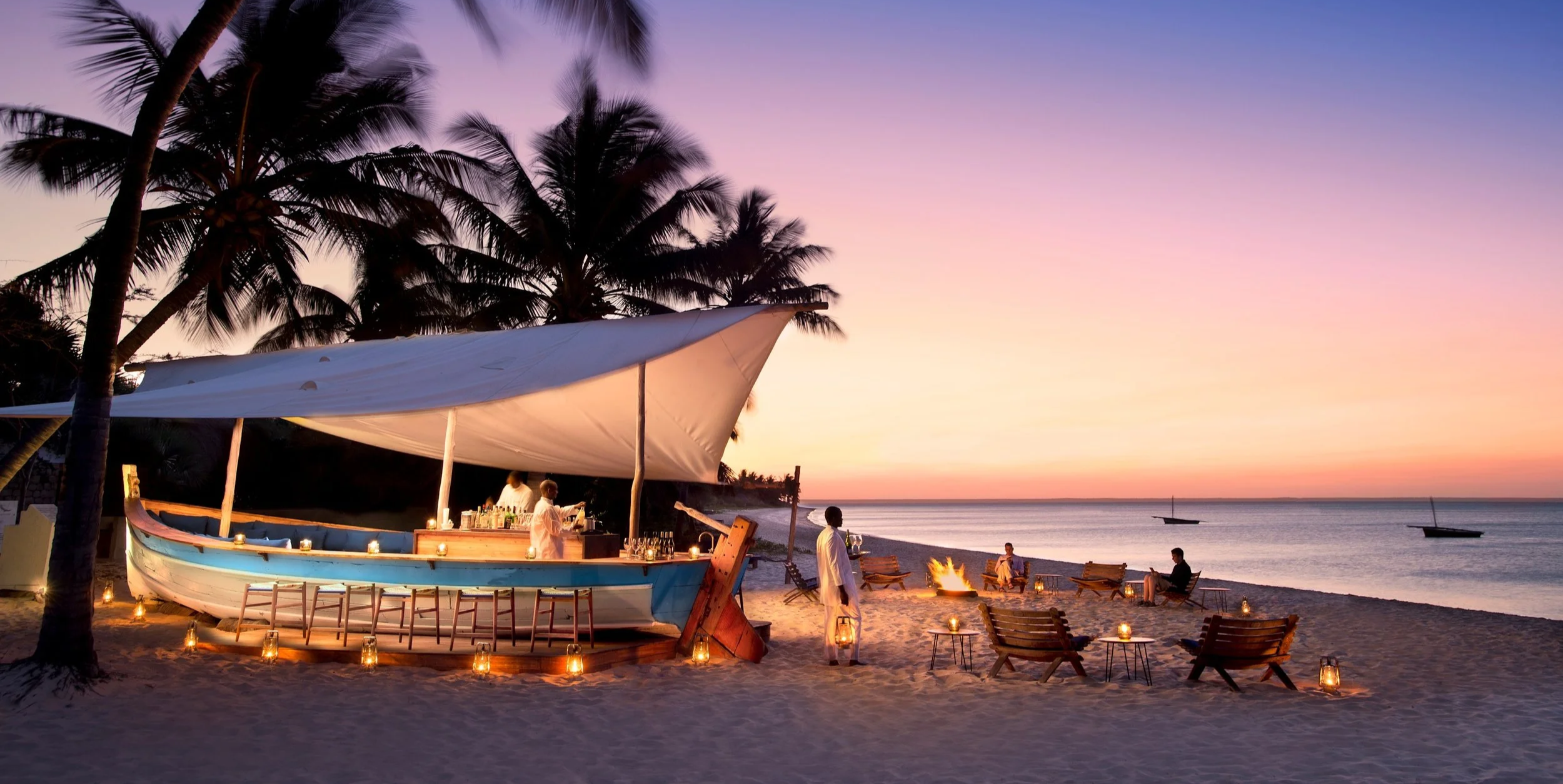 Beachside bar under a large canopy surrounded by palm trees, with candles lighting the sandy area. People are enjoying cocktail drinks by a fire on the beach during sunset, with boats floating on calm water in the background.