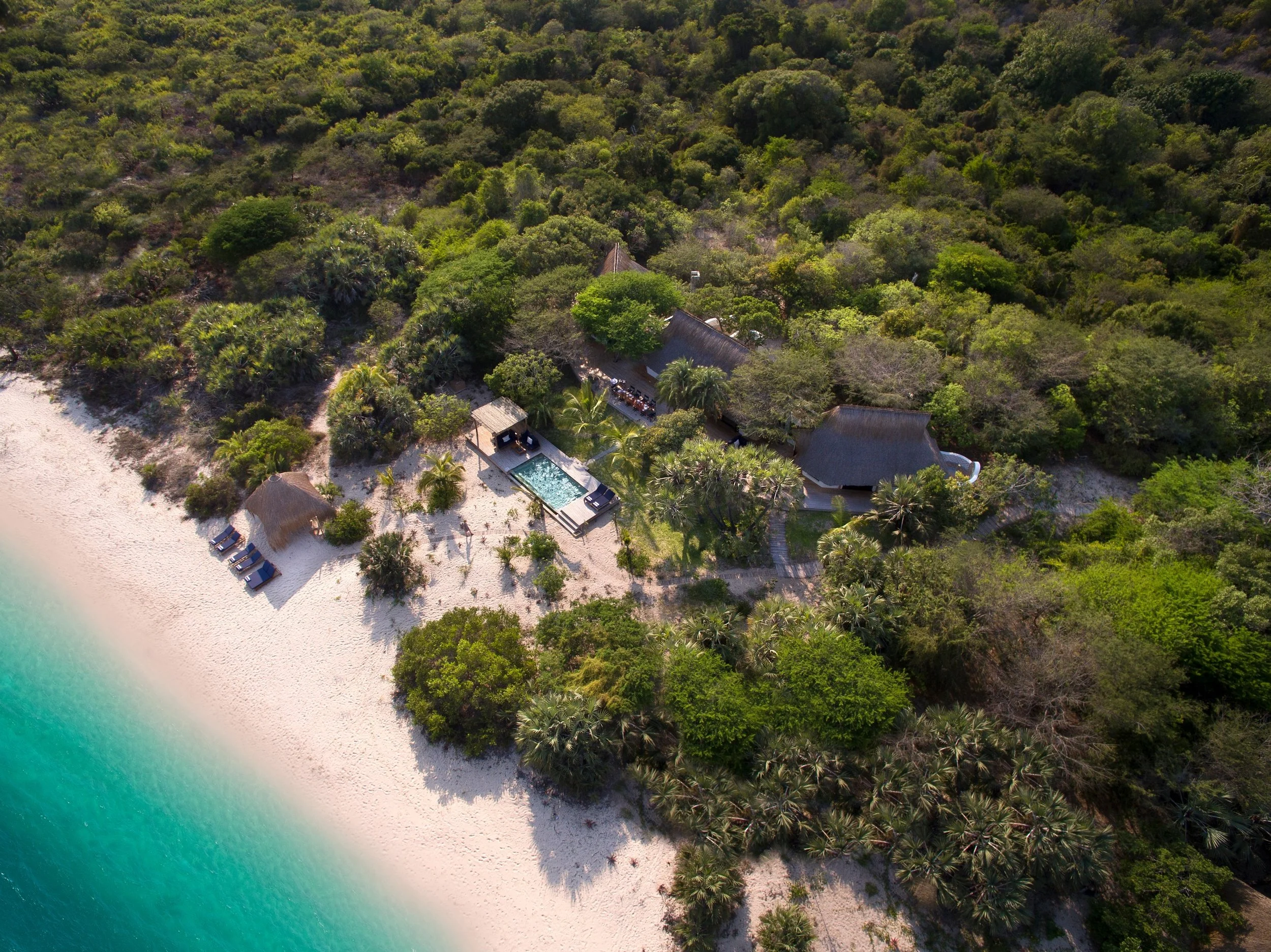 Aerial view of a tropical beach resort with white sand, turquoise water, and vegetation including palm trees. The resort has multiple thatched-roof structures, a swimming pool, and lounge chairs on the beach.