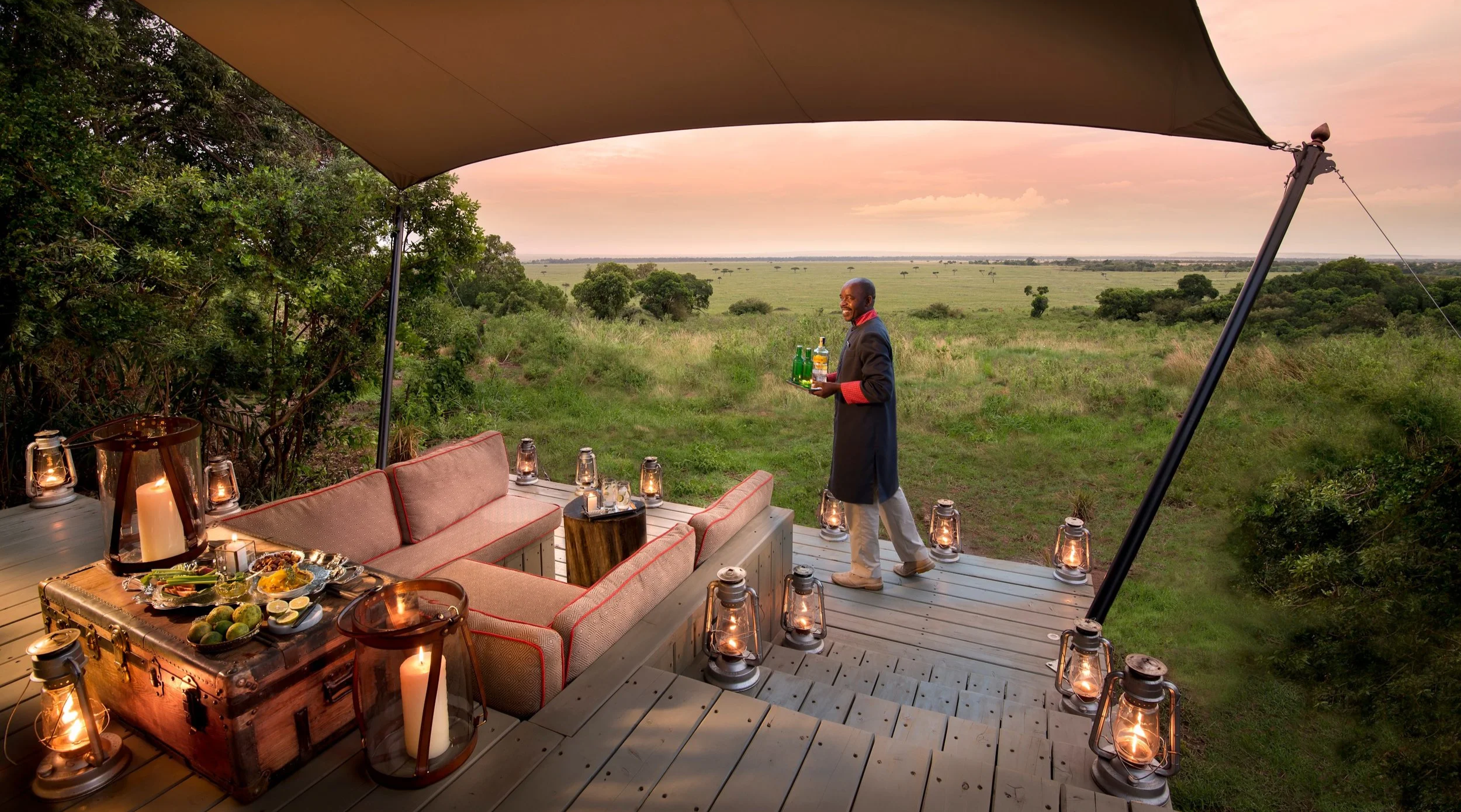 A waiter serving drinks on a wooden deck with outdoor furniture, surrounded by lanterns and candles, overlooking a lush open savannah landscape at sunset.