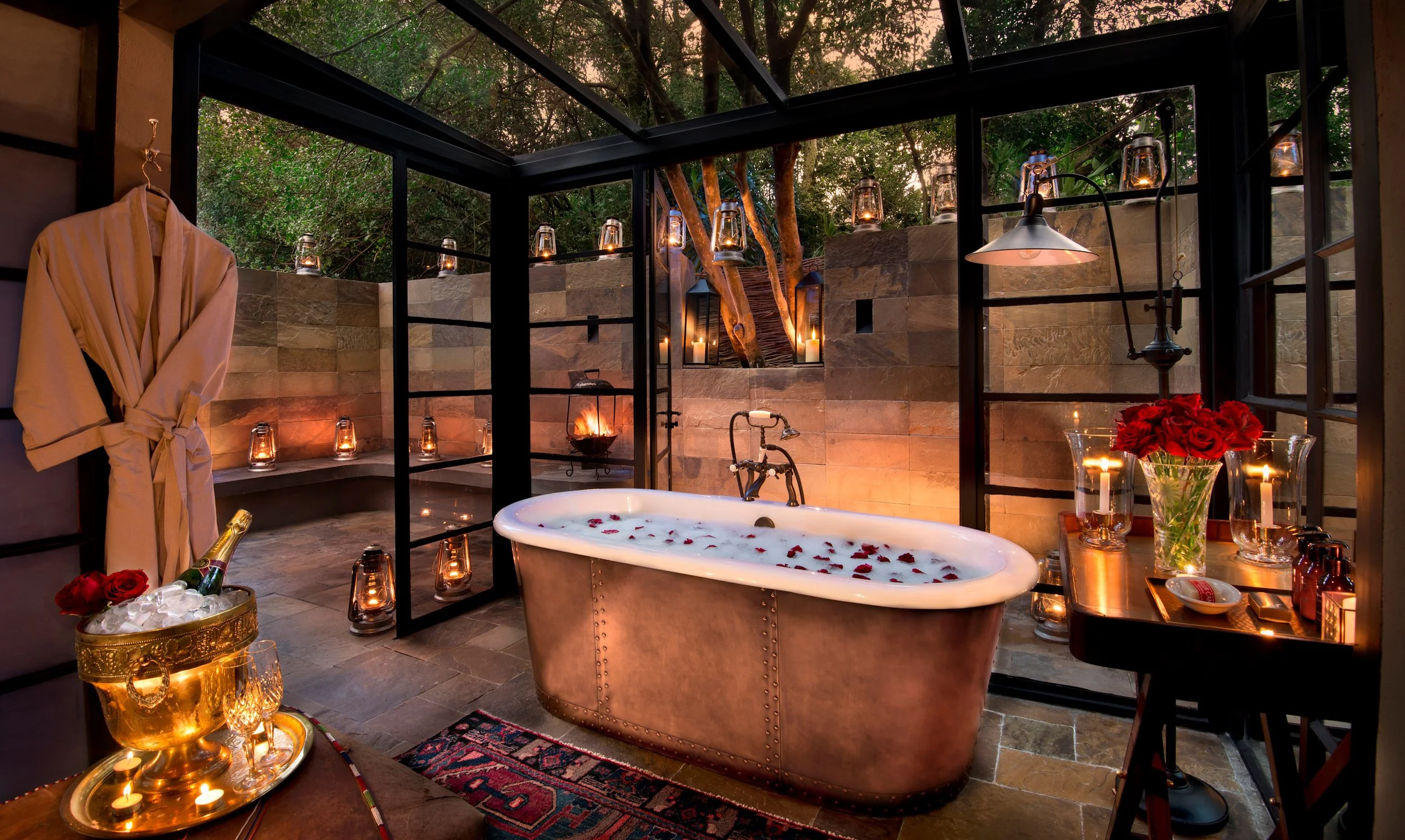 A luxurious indoor spa with a freestanding bathtub filled with water and rose petals, surrounded by candles, lanterns, and a bouquet of red roses on a side table, with a glass ceiling and lush green trees outside.