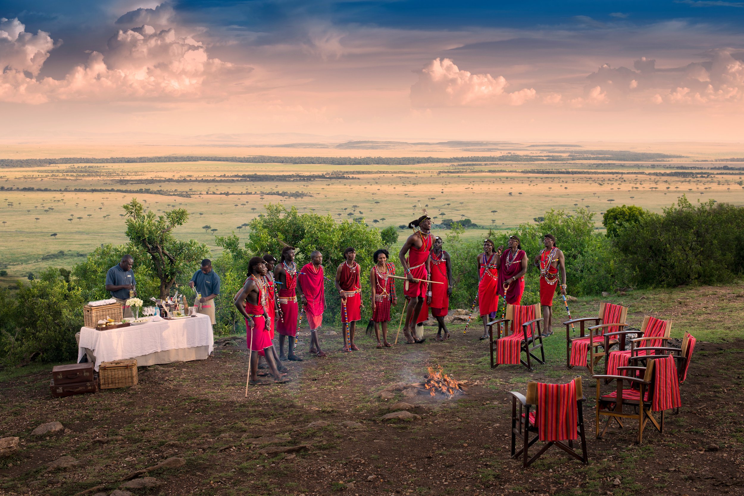 A group of Maasai people dressed in traditional red attire, some with jewelry and walking sticks, gathered around a small fire in an open landscape with a table of goods and chairs, under a cloudy sky.