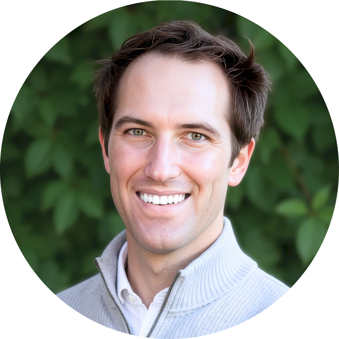 A smiling young man with brown hair and green eyes, wearing a light gray zip-up jacket over a white shirt, standing outdoors with green leaves in the background.