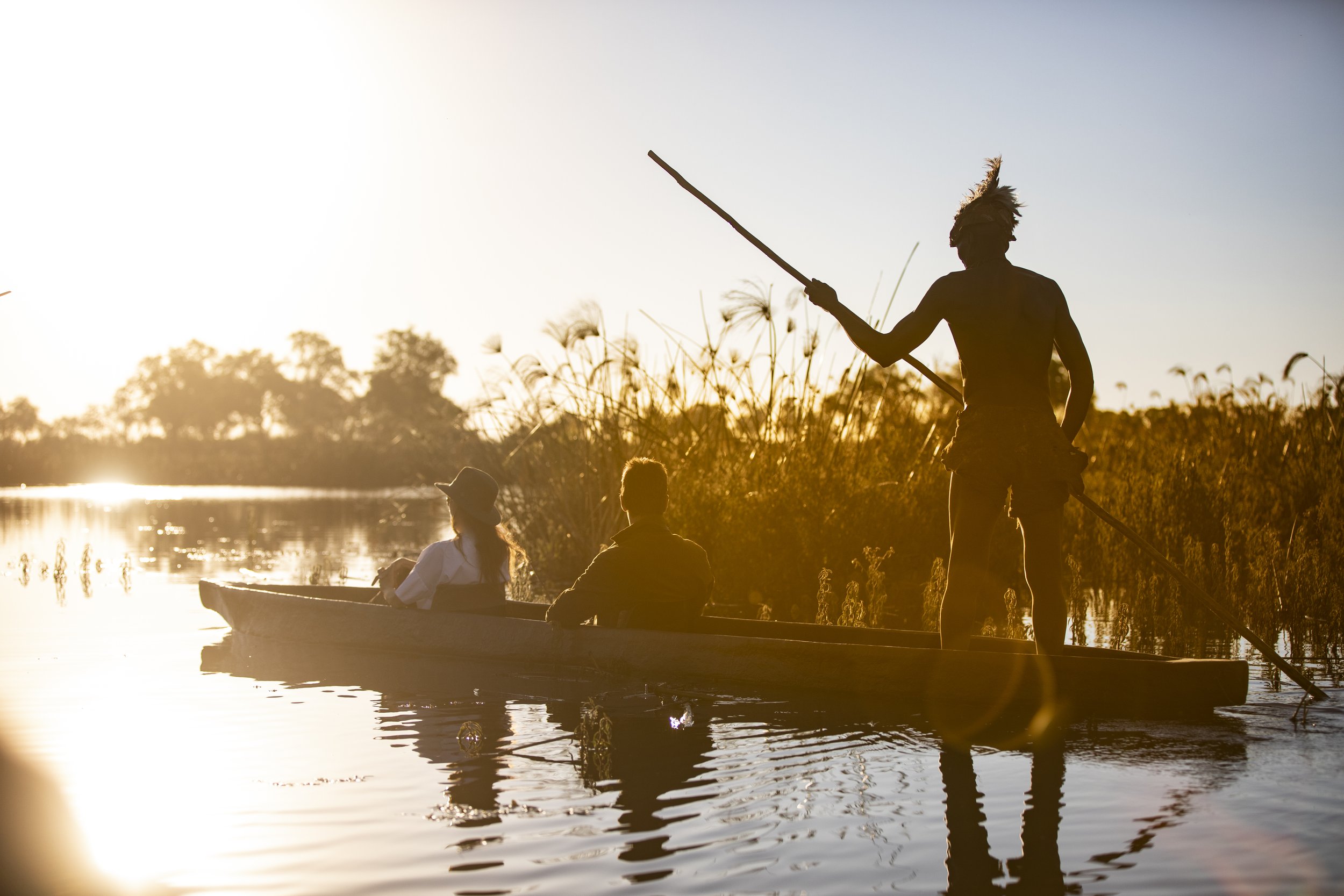 Silhouettes of three people on a boat and standing on the edge of a river during sunset, with tall grass and trees in the background.