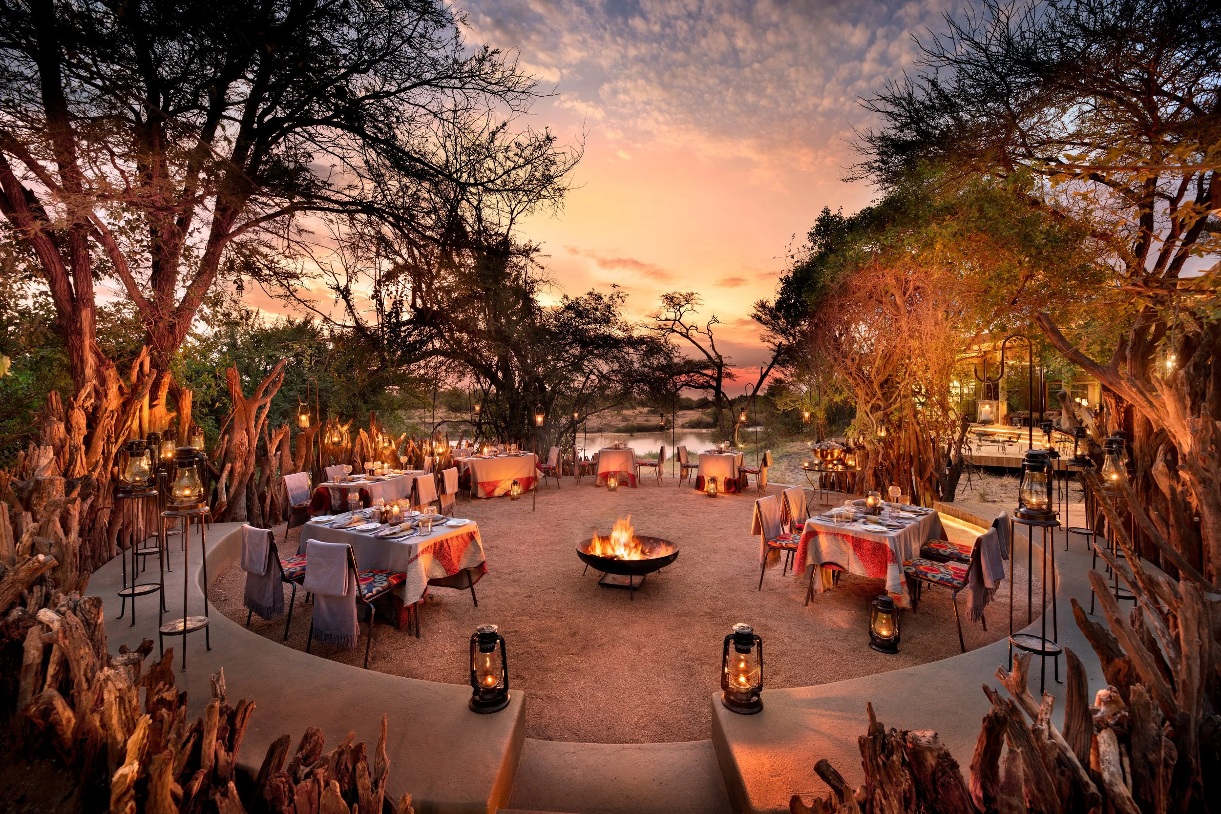 Outdoor dining area set up for a dinner in a natural setting during sunset, with tables, chairs, lanterns, and a fire pit in the center surrounded by trees.