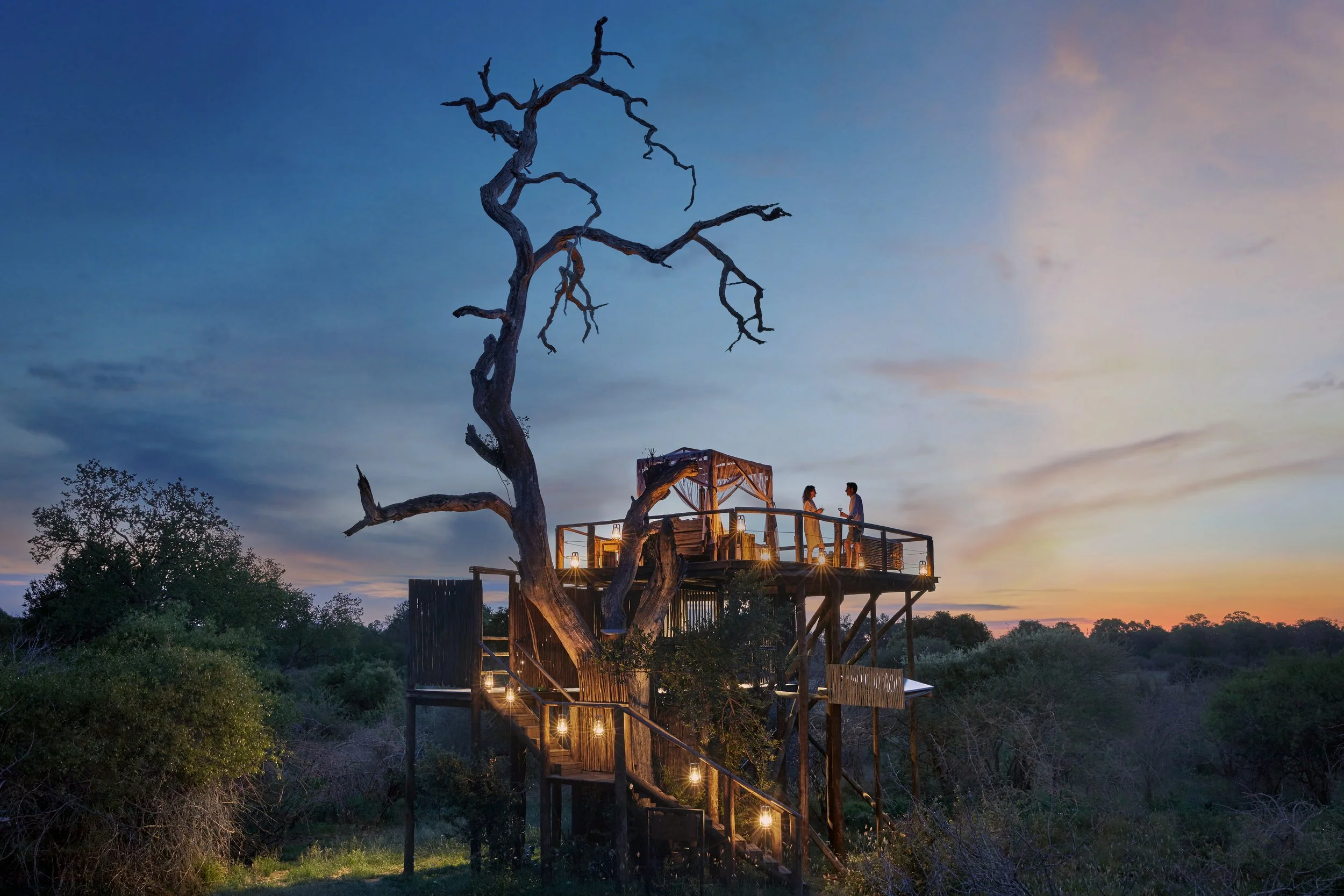 A treehouse structure built around a large, leafless tree at sunset, with two people standing on the balcony and warm string lights illuminating the stairs and balcony.