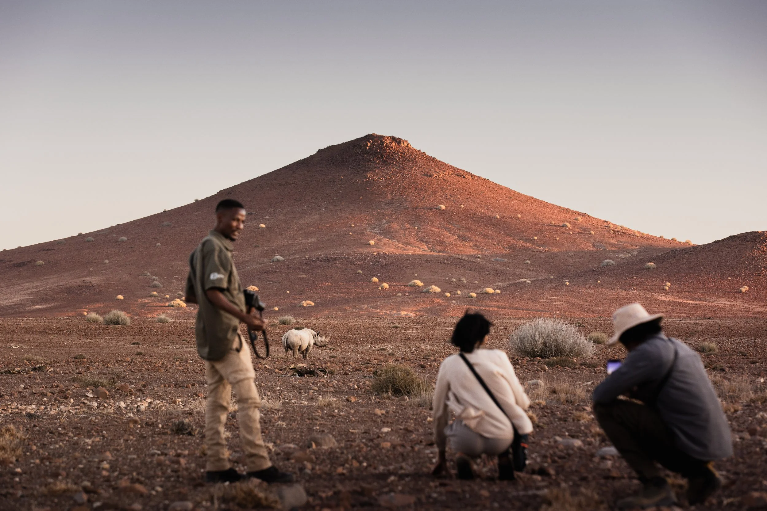 Three people in a desert landscape at sunset, one holding a camera, with a rhinoceros nearby and a large hill in the background.
