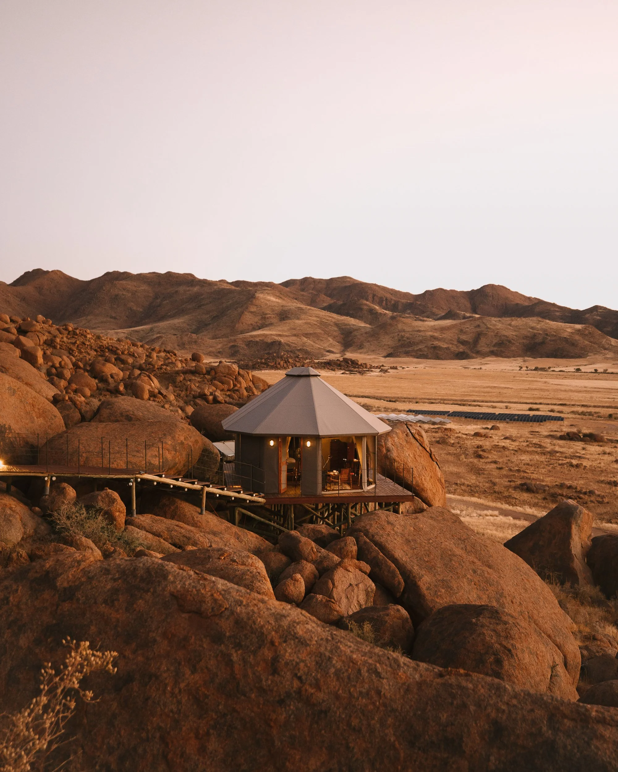 A round, modern house situated on a rocky hillside at sunset with mountains in the background.