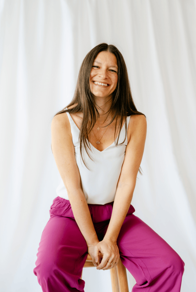 Junge Frau sitzt auf einem Hocker vor einem weißen Vorhang, lächelt und trägt weiße Top und pinke Hosen.