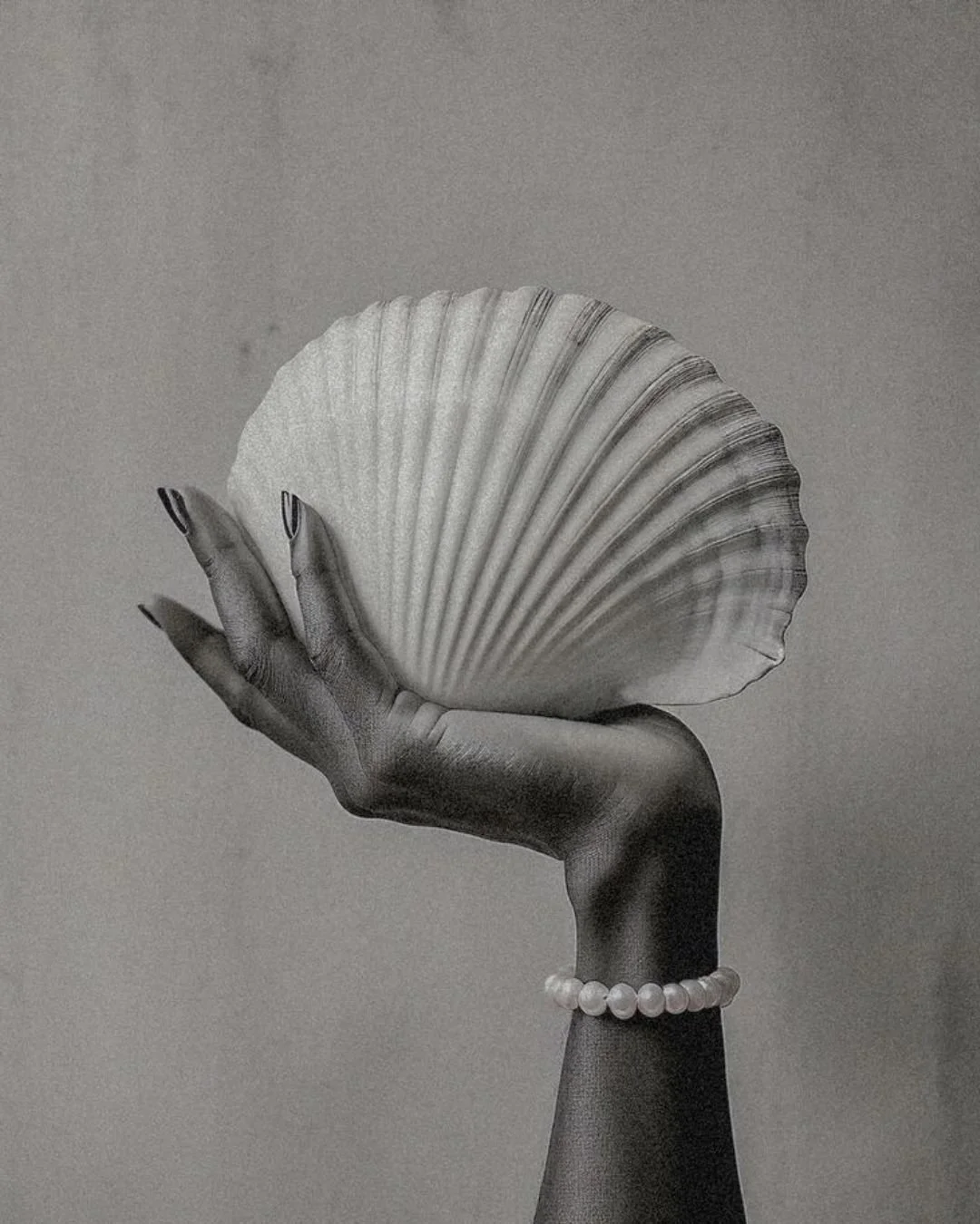 Schwarz-Weiß-Fotografie einer Hand, die eine große Muschel hält, mit Armband aus Perlen am Handgelenk.