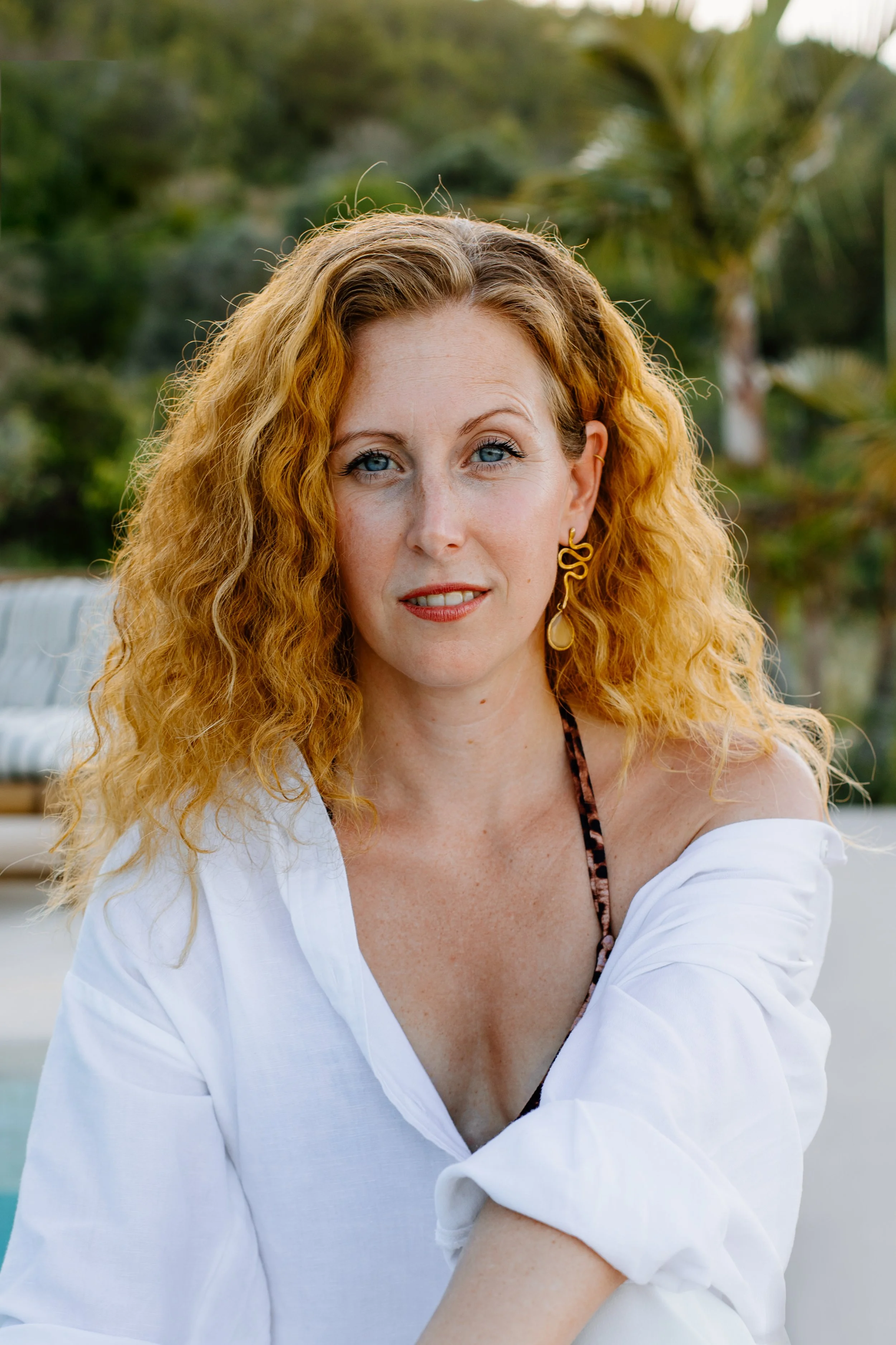 Nahaufnahme einer rotenhaaringen Frau mit blauen Augen, die eine weiße Bluse trägt, draußen im Garten, hinter ihr sind Bäume und Palmen.